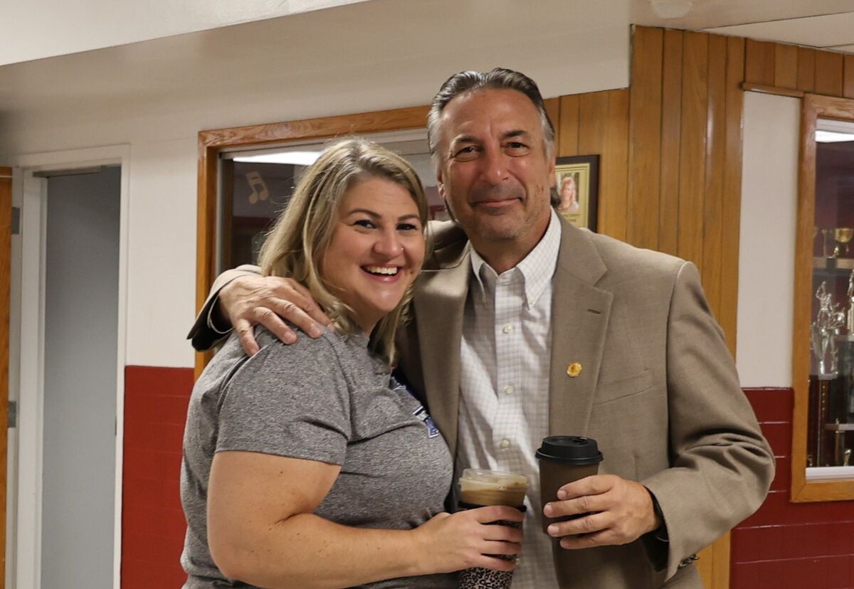 A smiling woman and a man in a blazer pose together indoors, each holding a beverage cup.