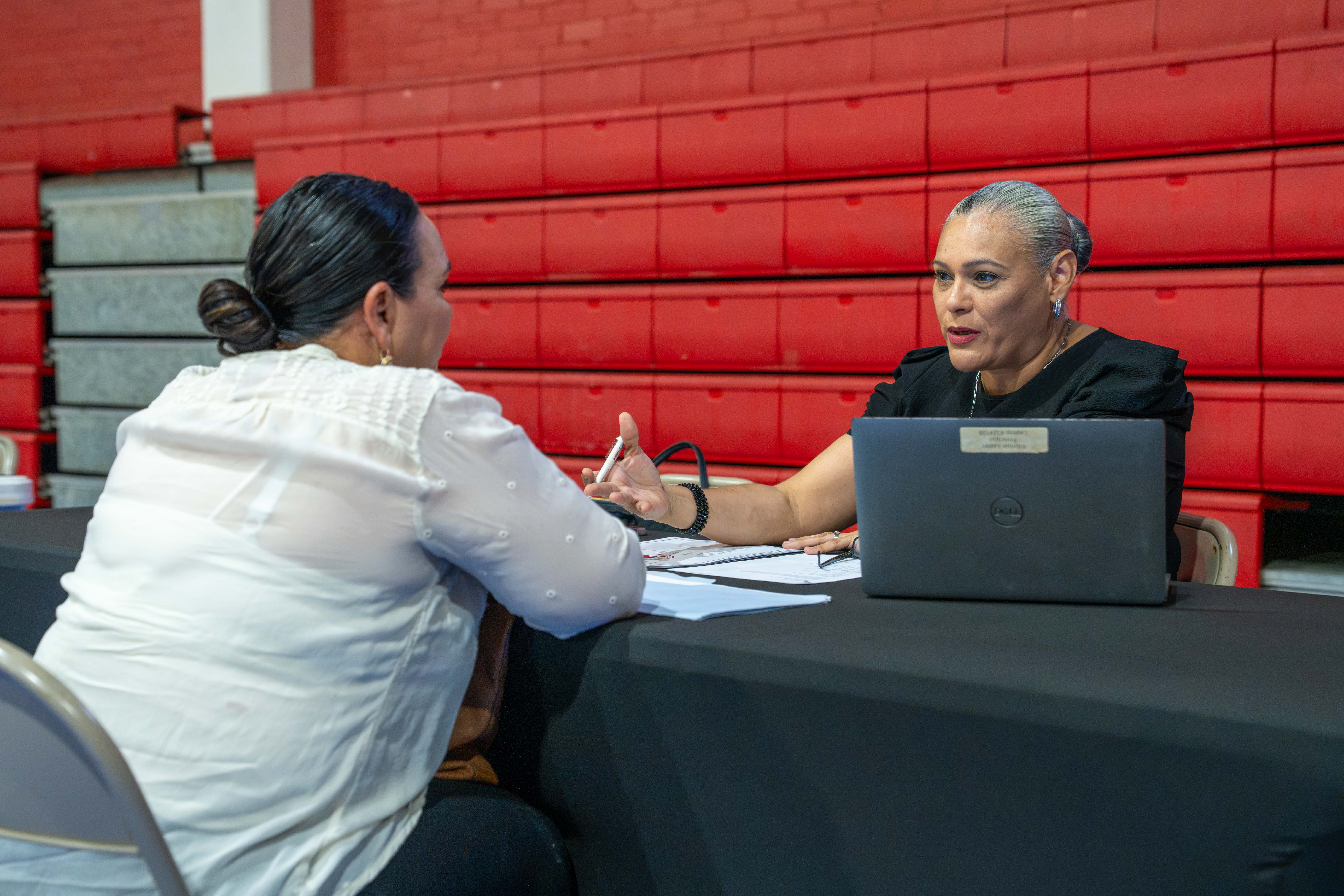 Two women are seated at a table, one holding a pen and gesturing while speaking to the other, who is looking at papers.