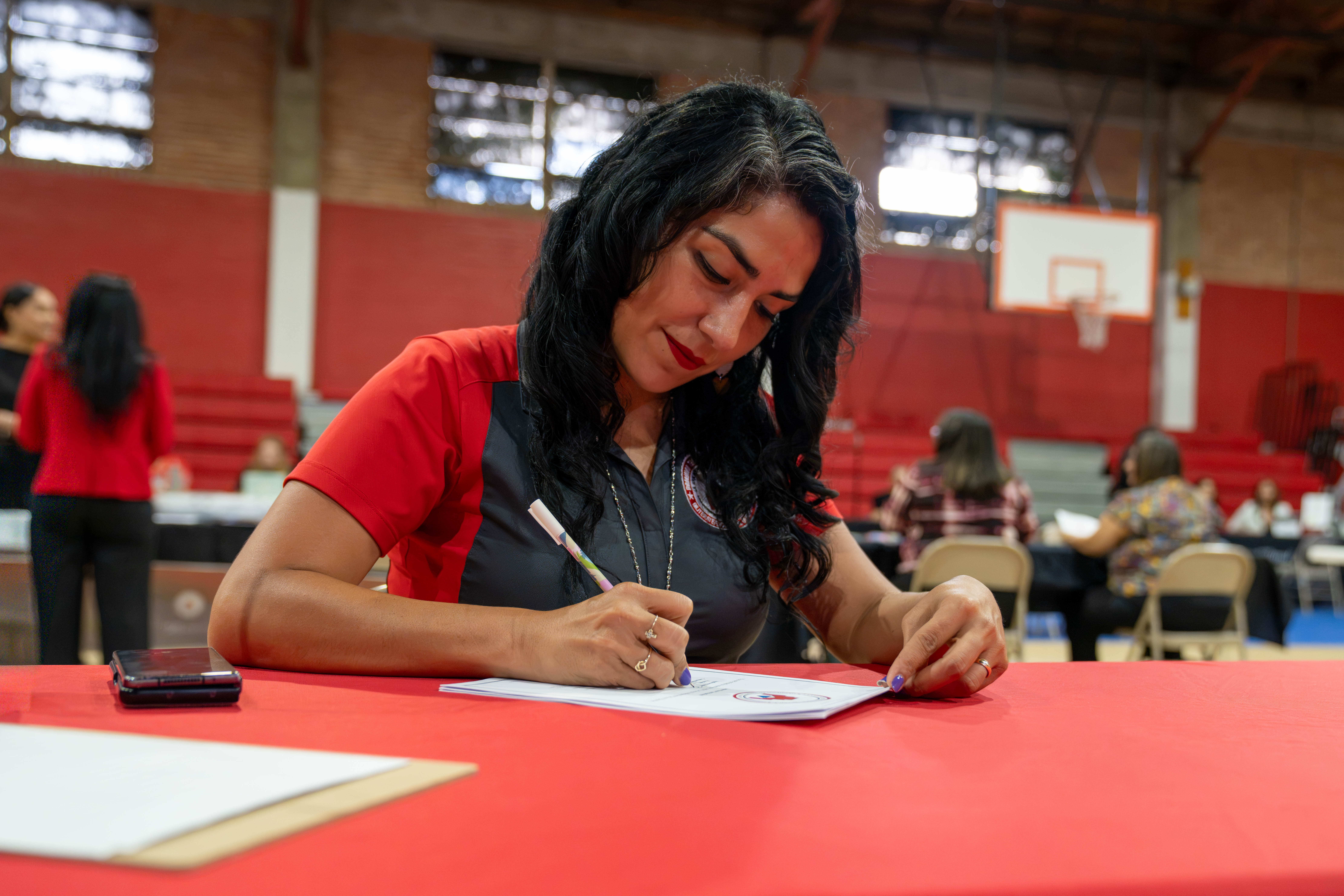 A woman with dark hair and red lipstick is signing a document at a red table in a gymnasium.
