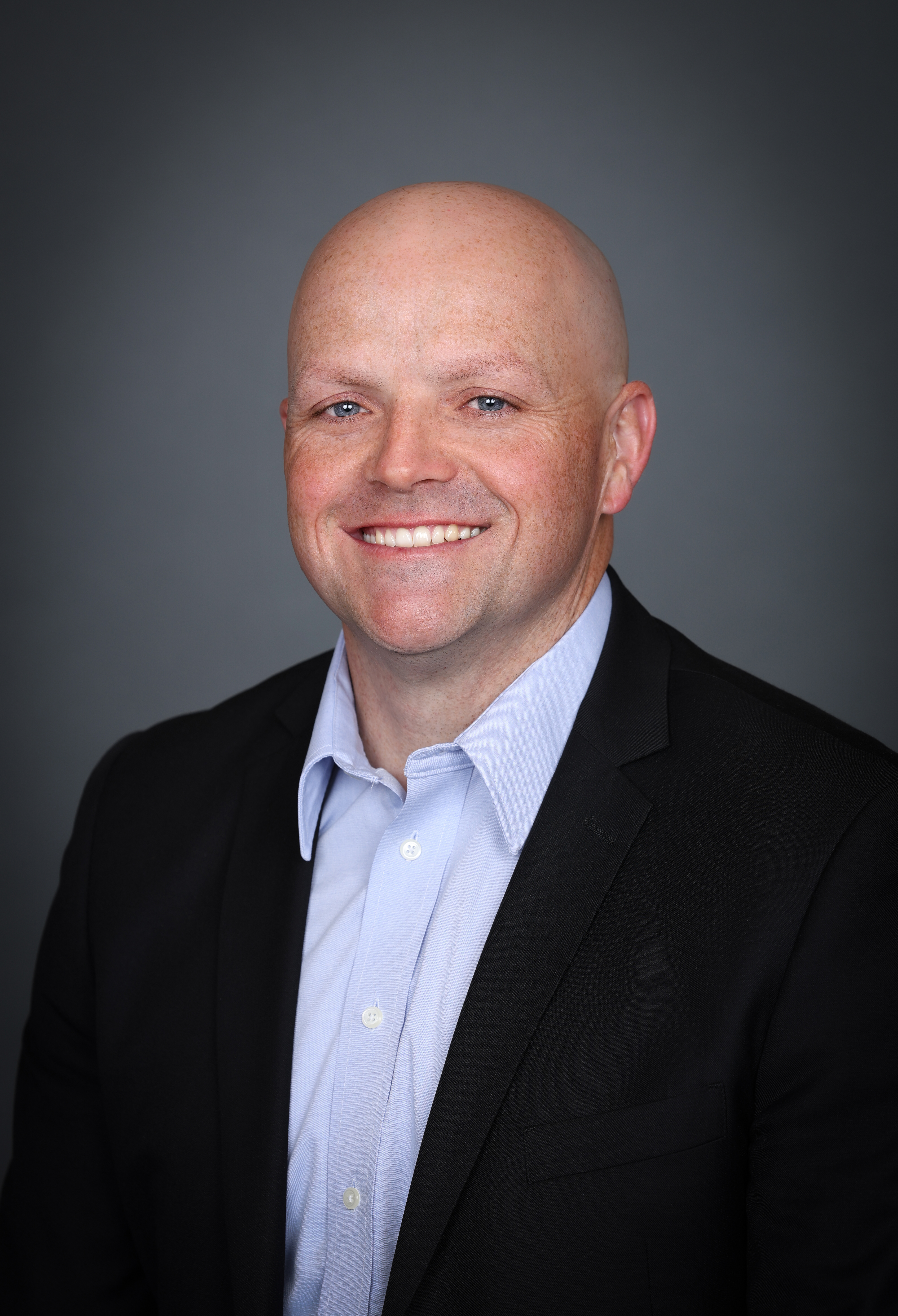 A professional headshot of a smiling man with a shaved head, wearing a light blue button-down shirt and a black blazer against a grey studio background.