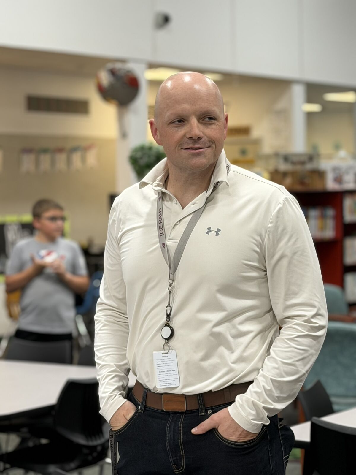 A bald man in a white long-sleeved shirt and lanyard smiles while standing in a brightly lit indoor space.