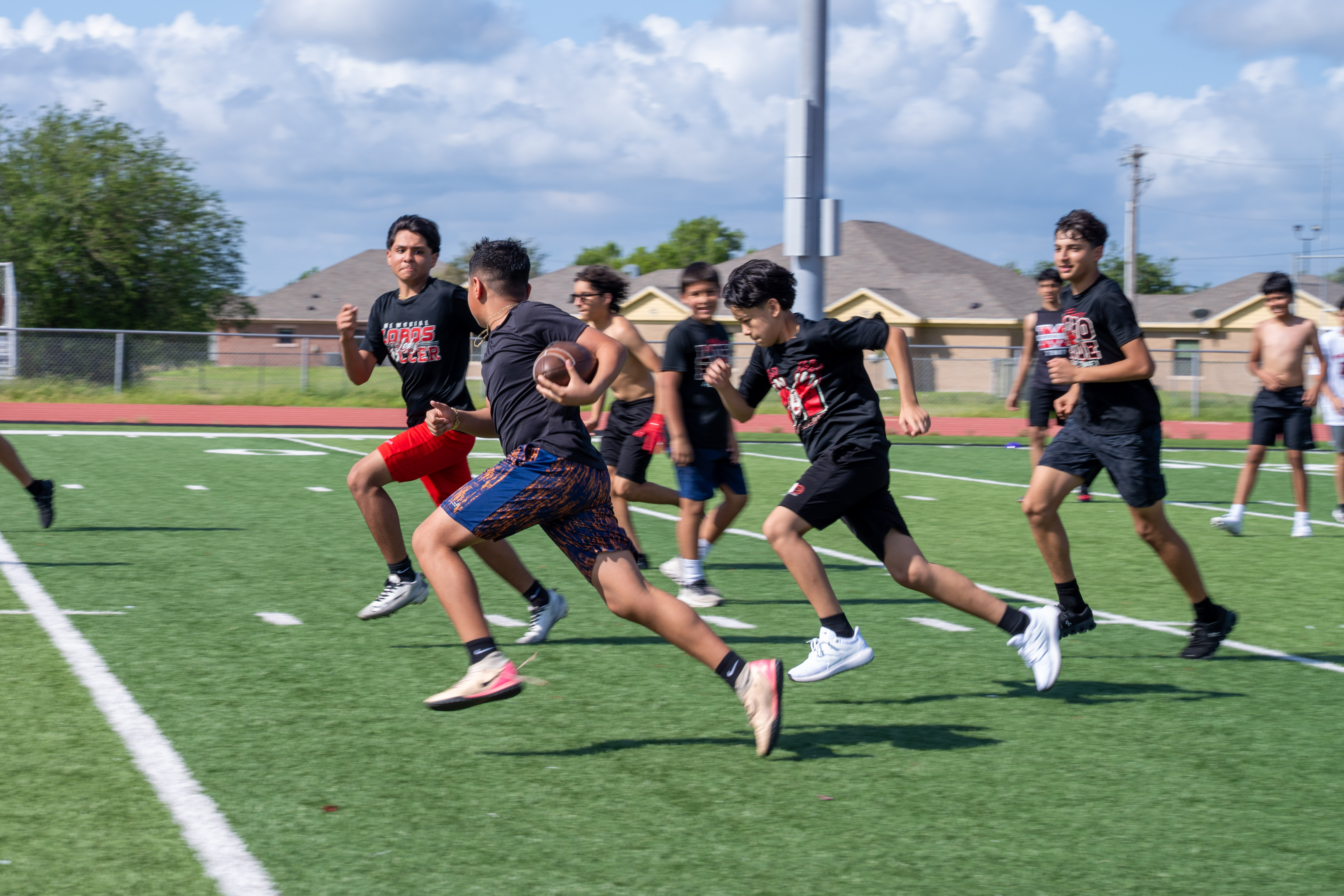 A group of young athletes practice on a green turf field under a bright sky.