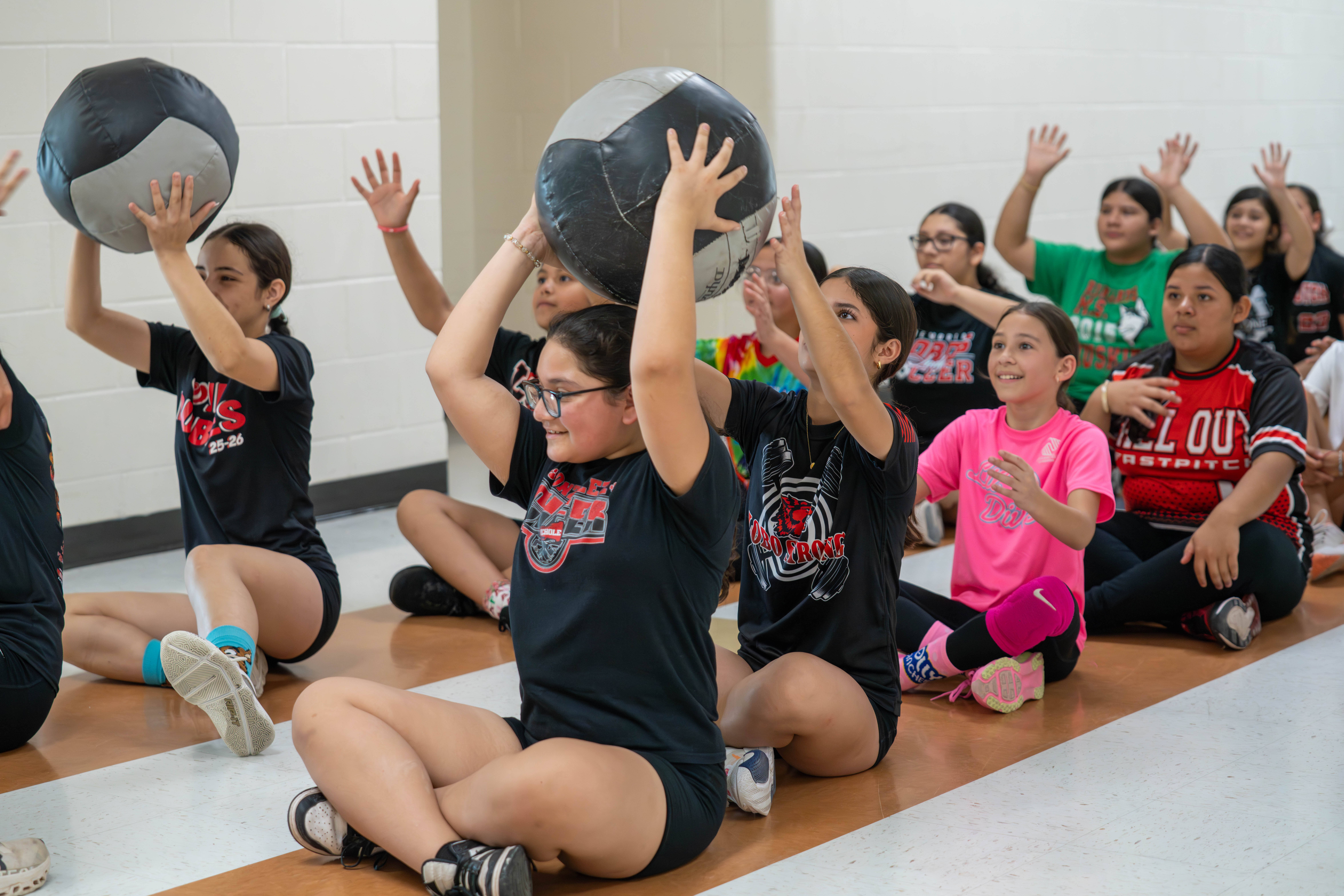 A group of young people sit on the floor, holding a large medicine ball during an exercise class.
