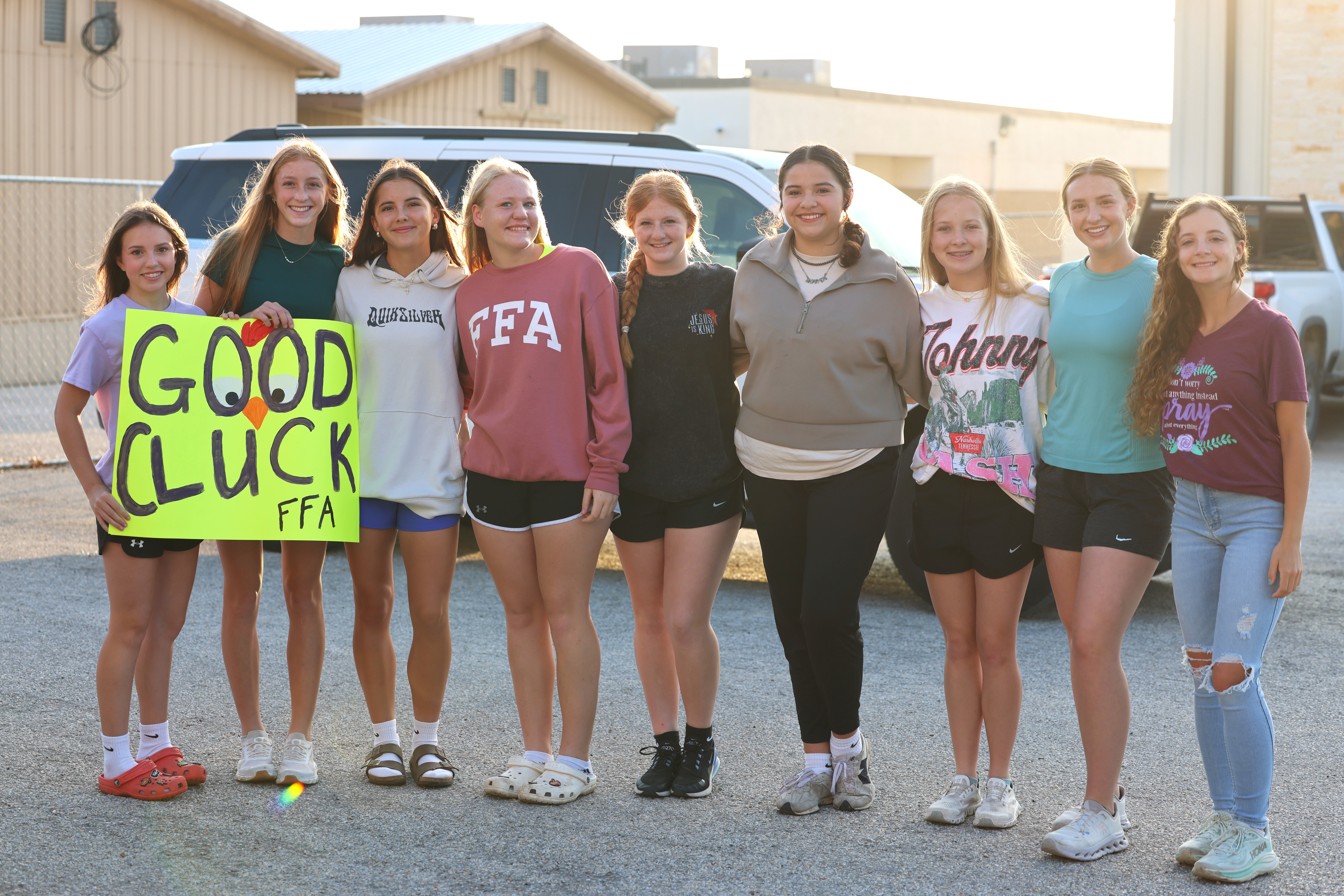A group of nine young women stand together outdoors, with one holding a bright yellow sign that reads 'GOOD CLUCK FFA'.