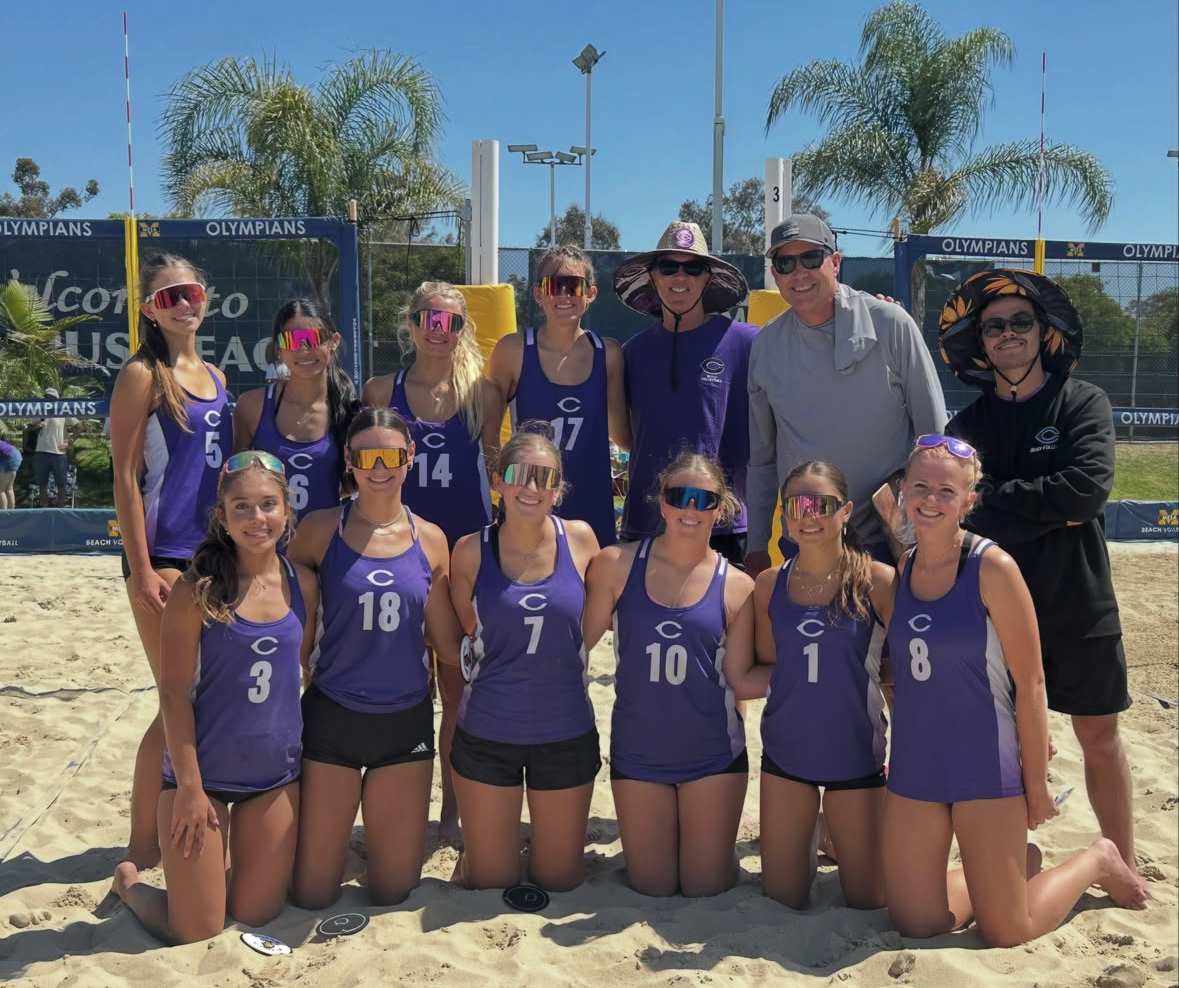 A group of young women in purple volleyball uniforms pose for a photo on a sandy beach court.
