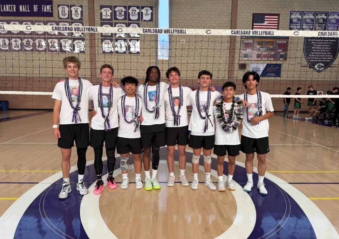 Eight young men in white t-shirts and black shorts stand together on a volleyball court, wearing leis with photos.