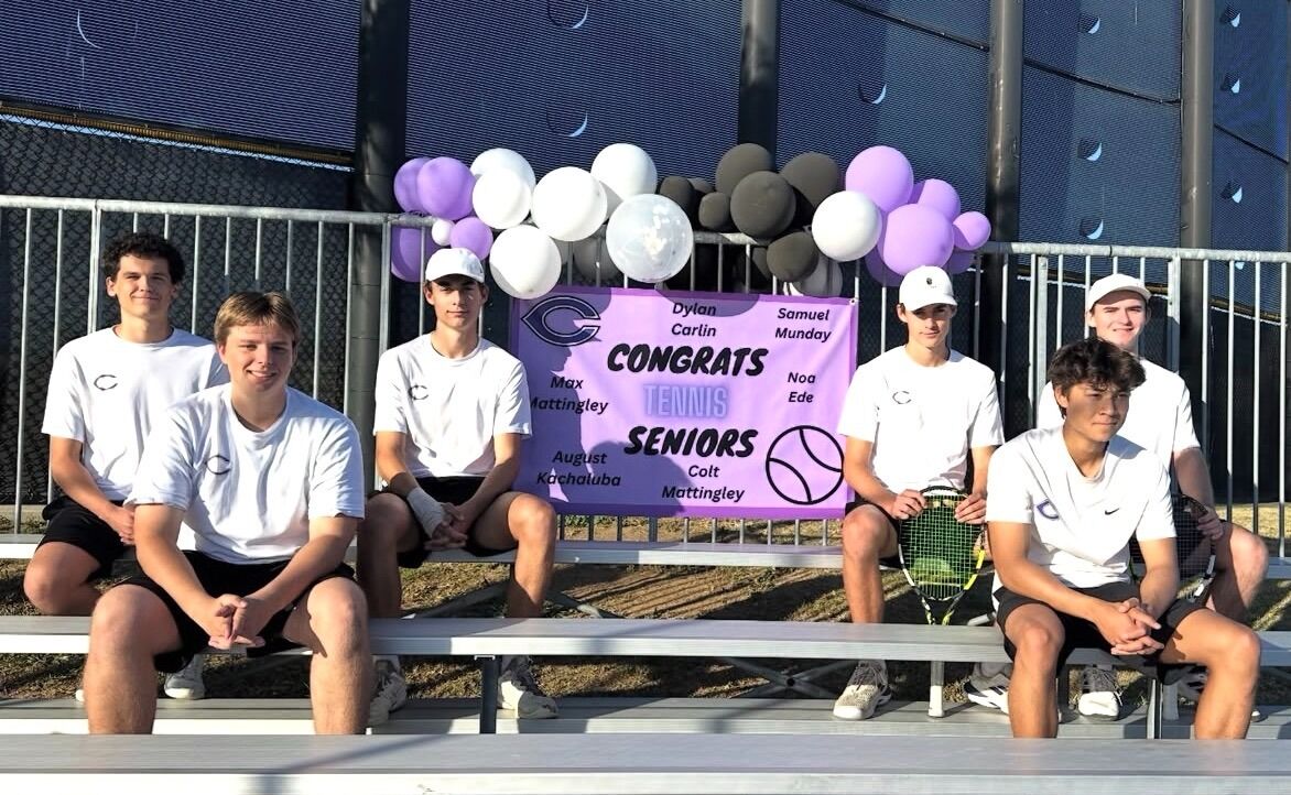 Six young men in tennis attire sit on bleachers, celebrating with a banner that reads 'CONGRATS TENNIS SENIORS'.