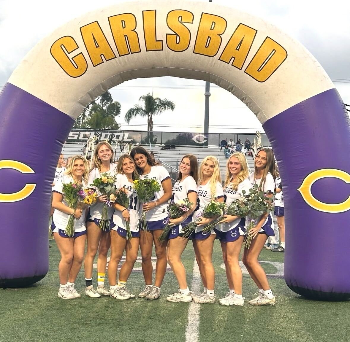 Seven young women in athletic uniforms stand under a large inflatable arch with 'CARLSBAD' written across it.