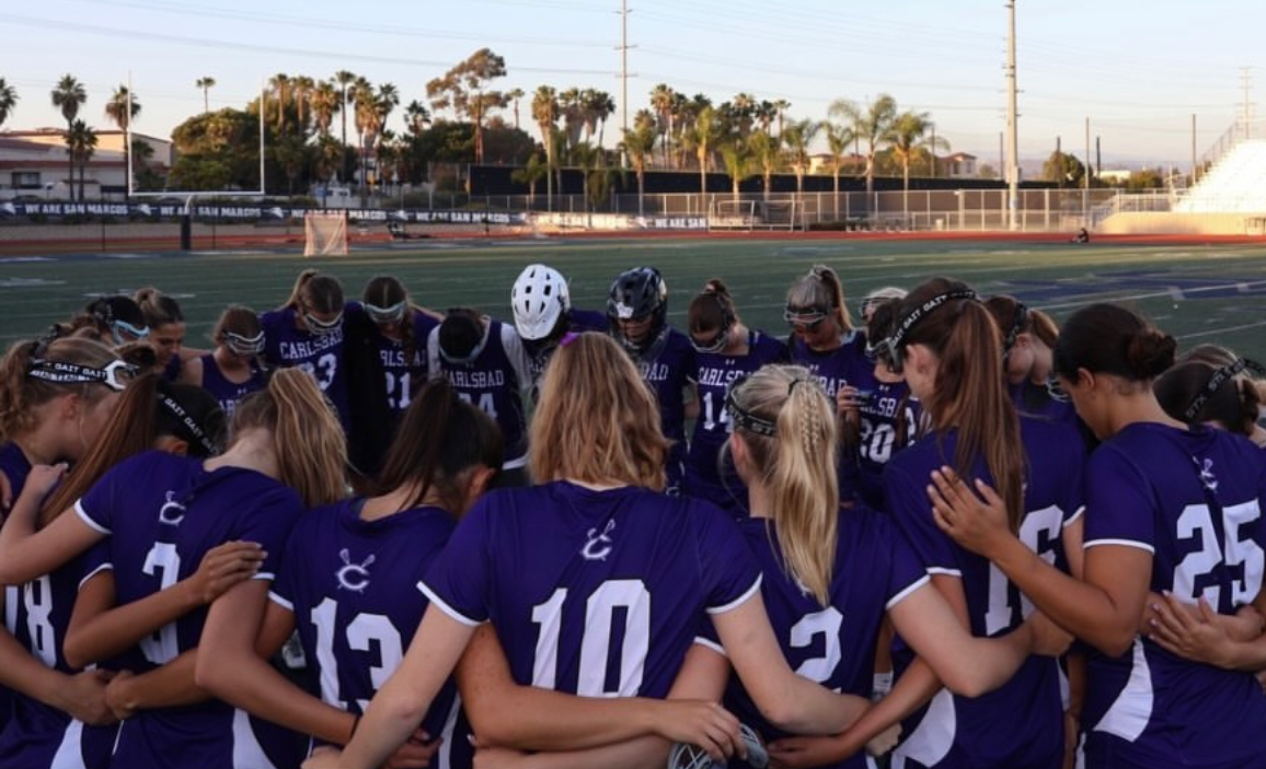 A girls' lacrosse team in purple uniforms huddles together on a field, with their heads bowed.