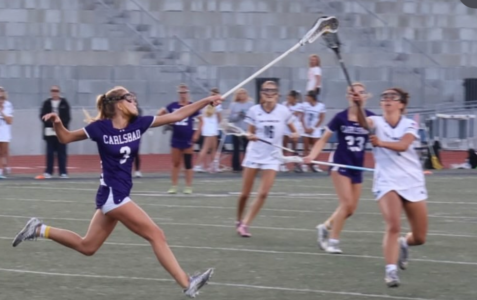 A lacrosse player in a purple uniform with 'CARLSBAD' on it reaches for the ball.