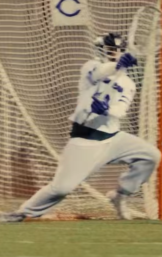 A lacrosse goalie in a white and blue uniform dives to make a save in front of the net.