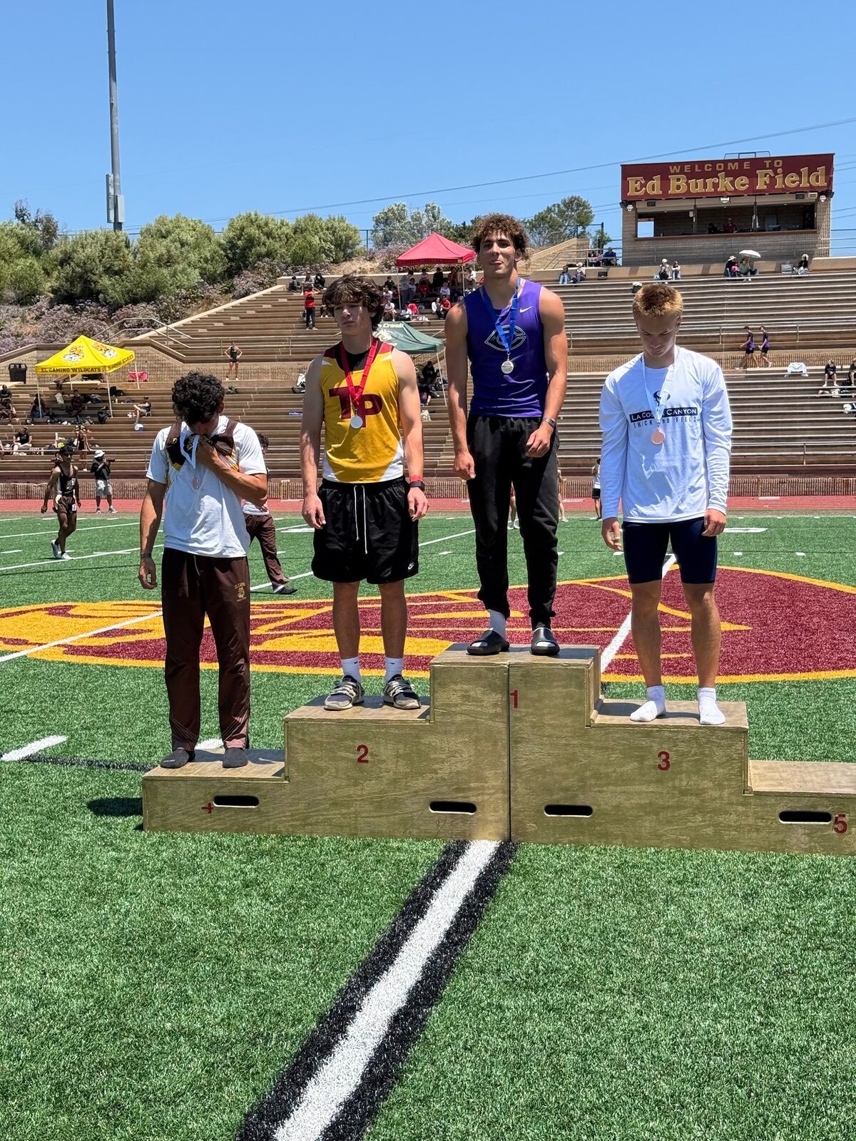 Four young athletes stand on a wooden podium on a sunny day at Ed Burke Field.