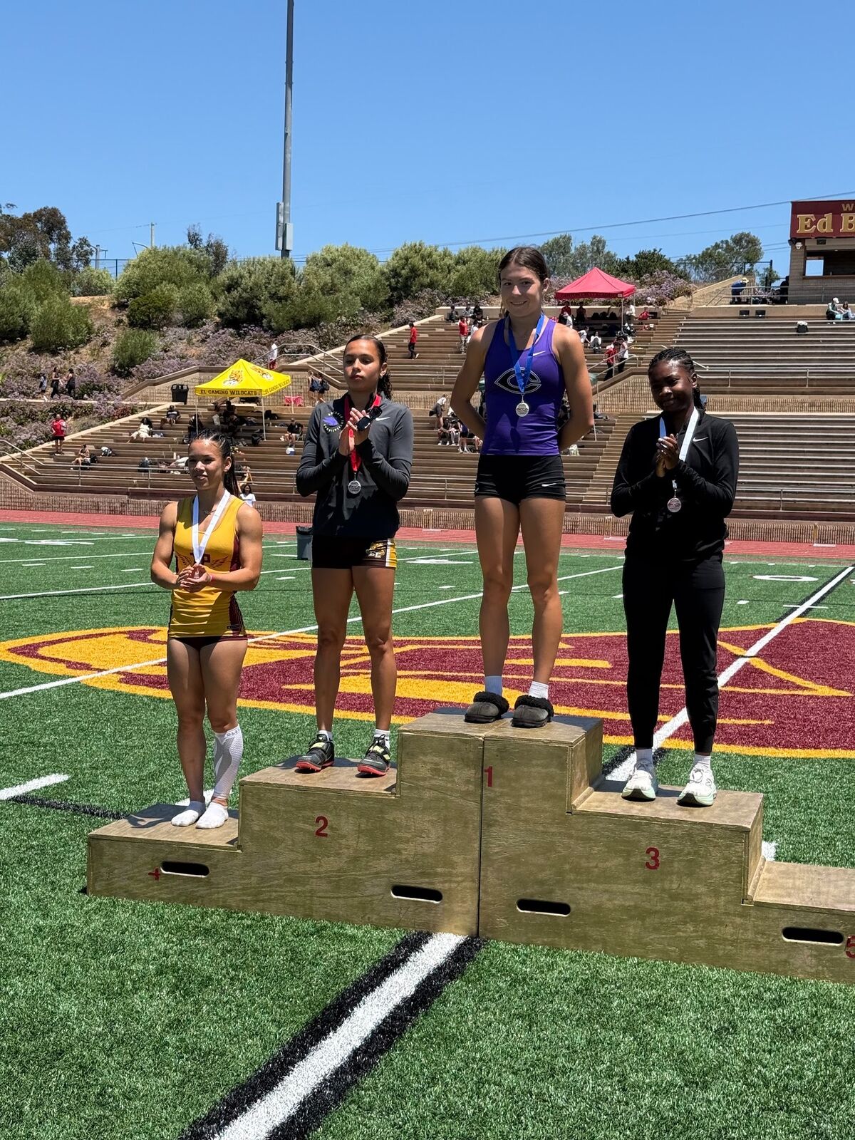 Four female athletes stand on a wooden awards podium on a sunny day at a track and field event.