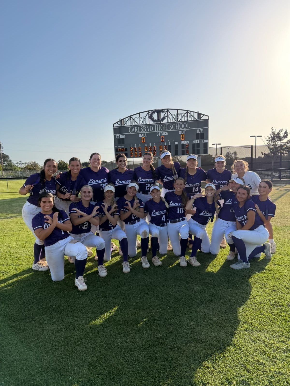 A softball team poses for a photo on a grassy field in front of a scoreboard at Carlsbad High School.