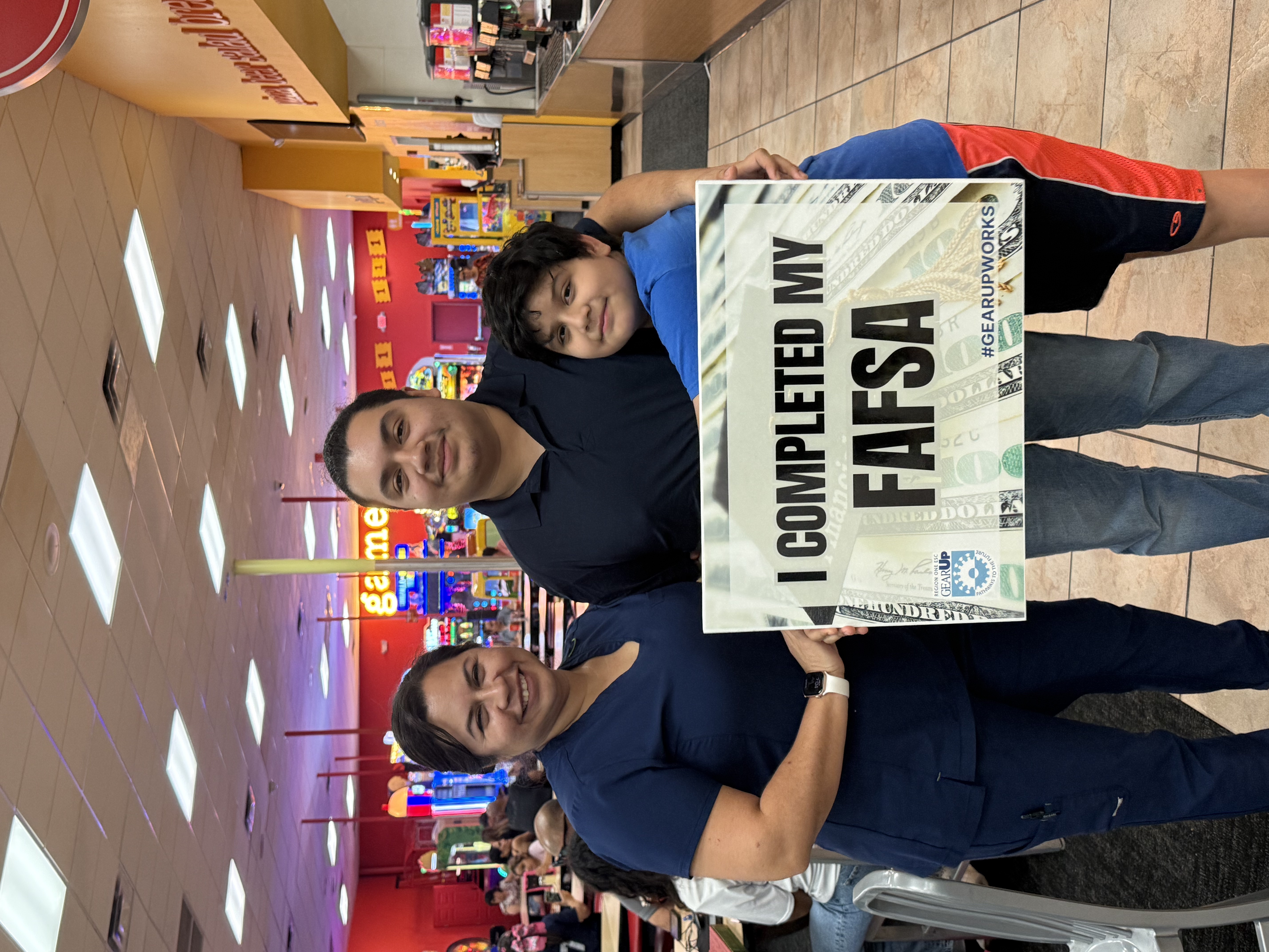 A family holds a sign that reads "I COMPLETED MY FAFSA" inside a brightly lit restaurant or arcade setting.