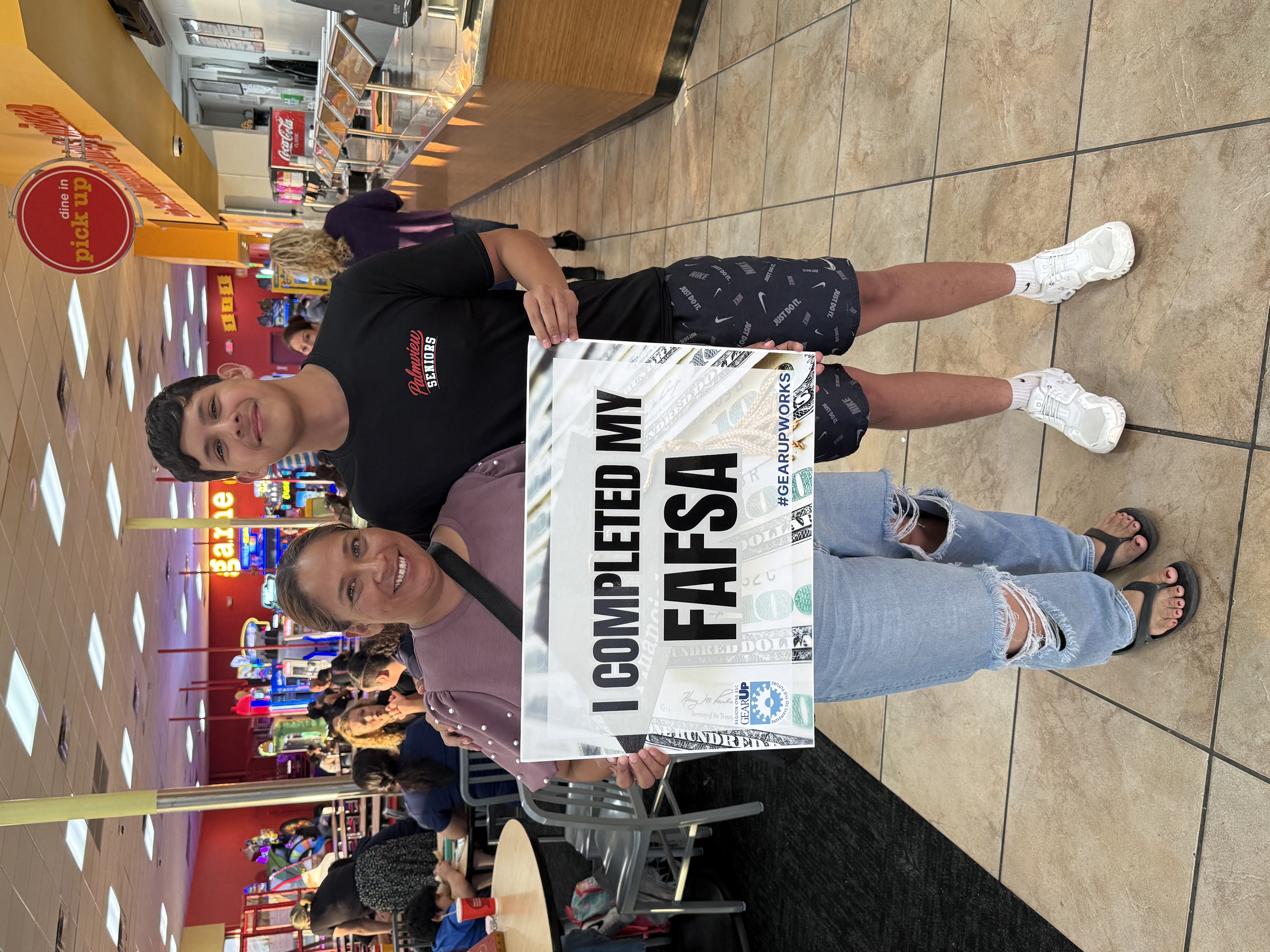 A smiling teenager and adult hold a sign that reads, "I COMPLETED MY FAFSA," standing indoors in a public area.