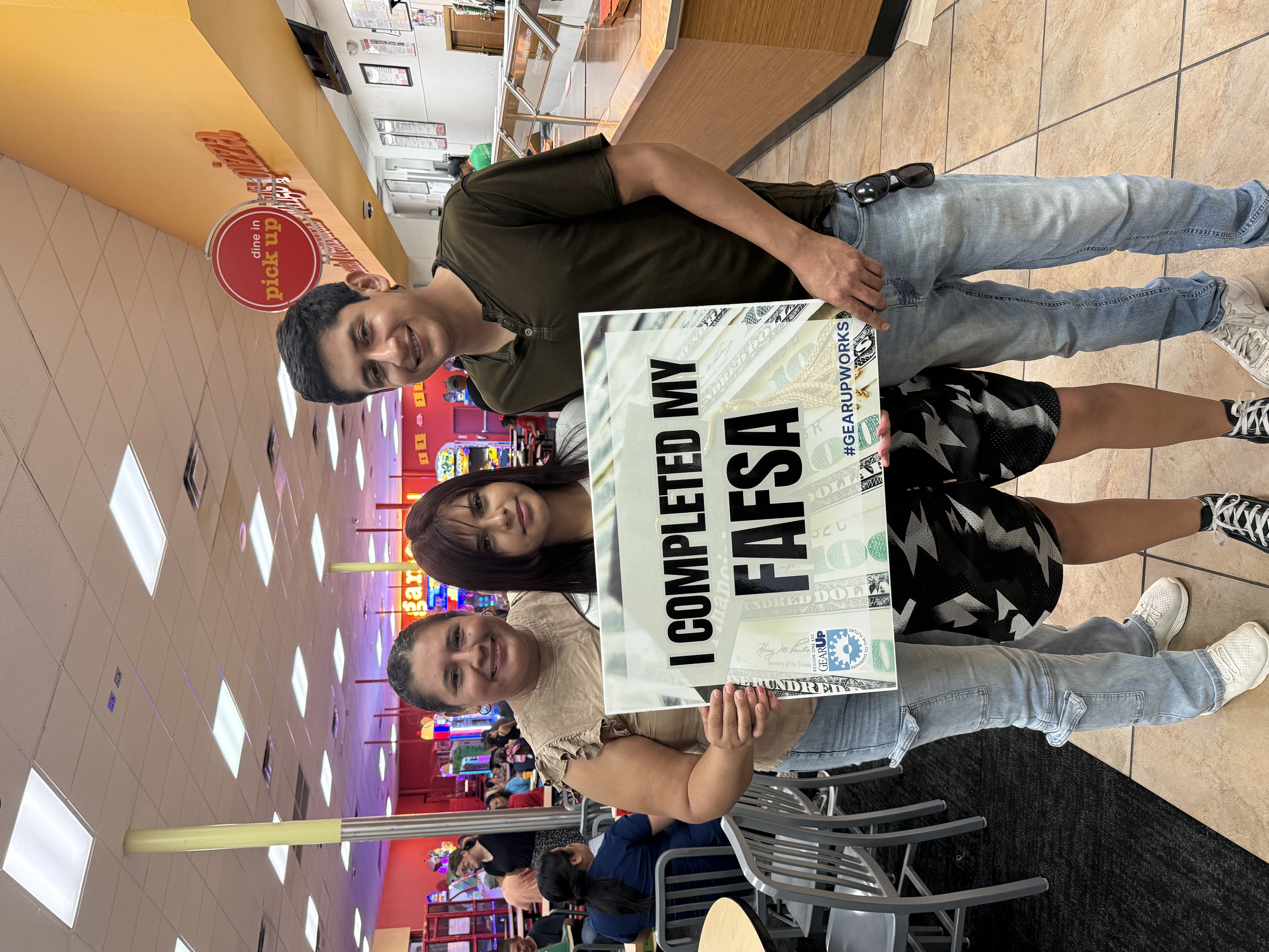 Three people proudly hold a sign that reads "I COMPLETED MY FAFSA" inside a brightly lit restaurant or cafeteria.
