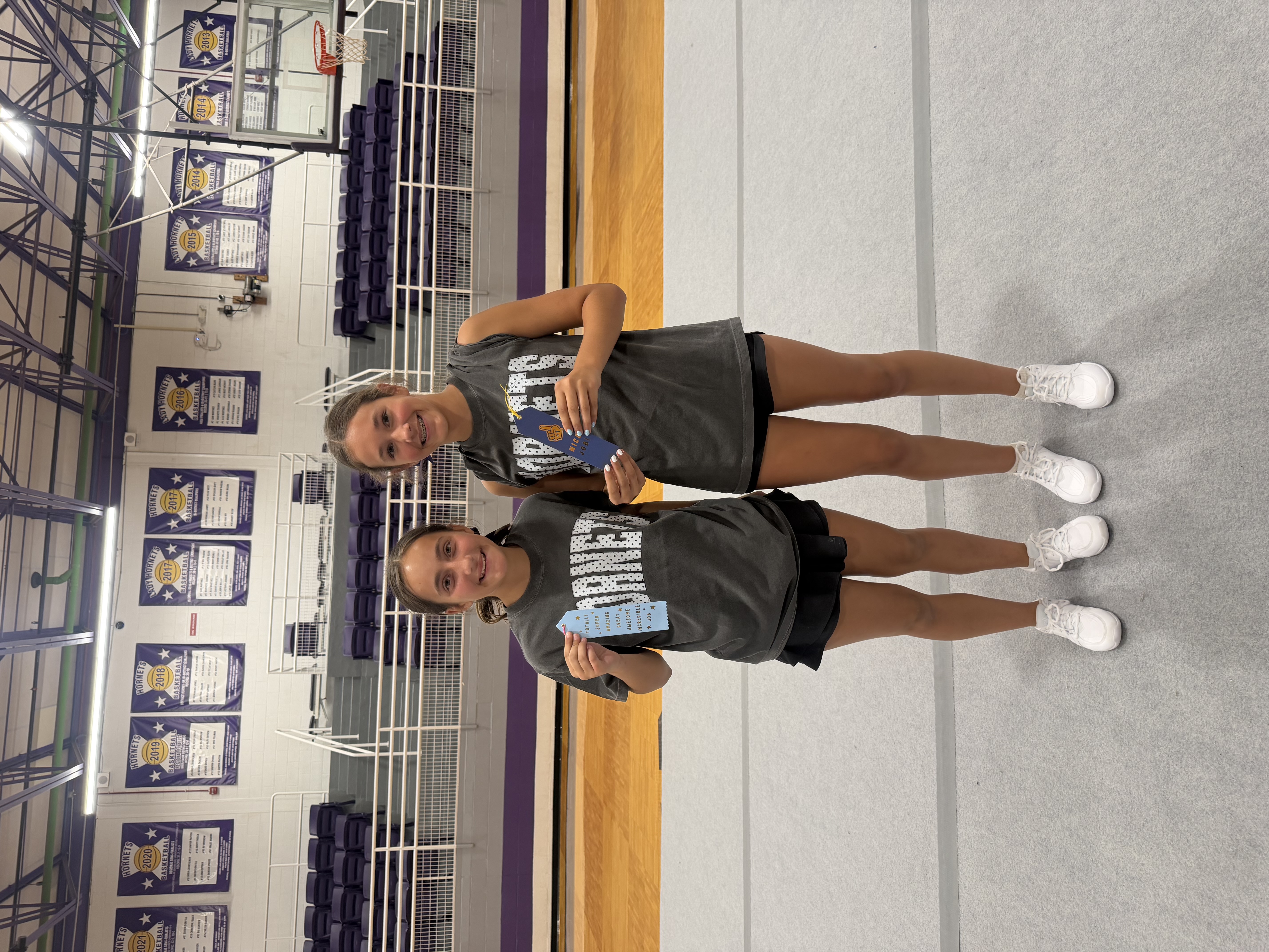 Two young girls in matching grey t-shirts smile, each holding a blue ribbon award in a gymnasium.