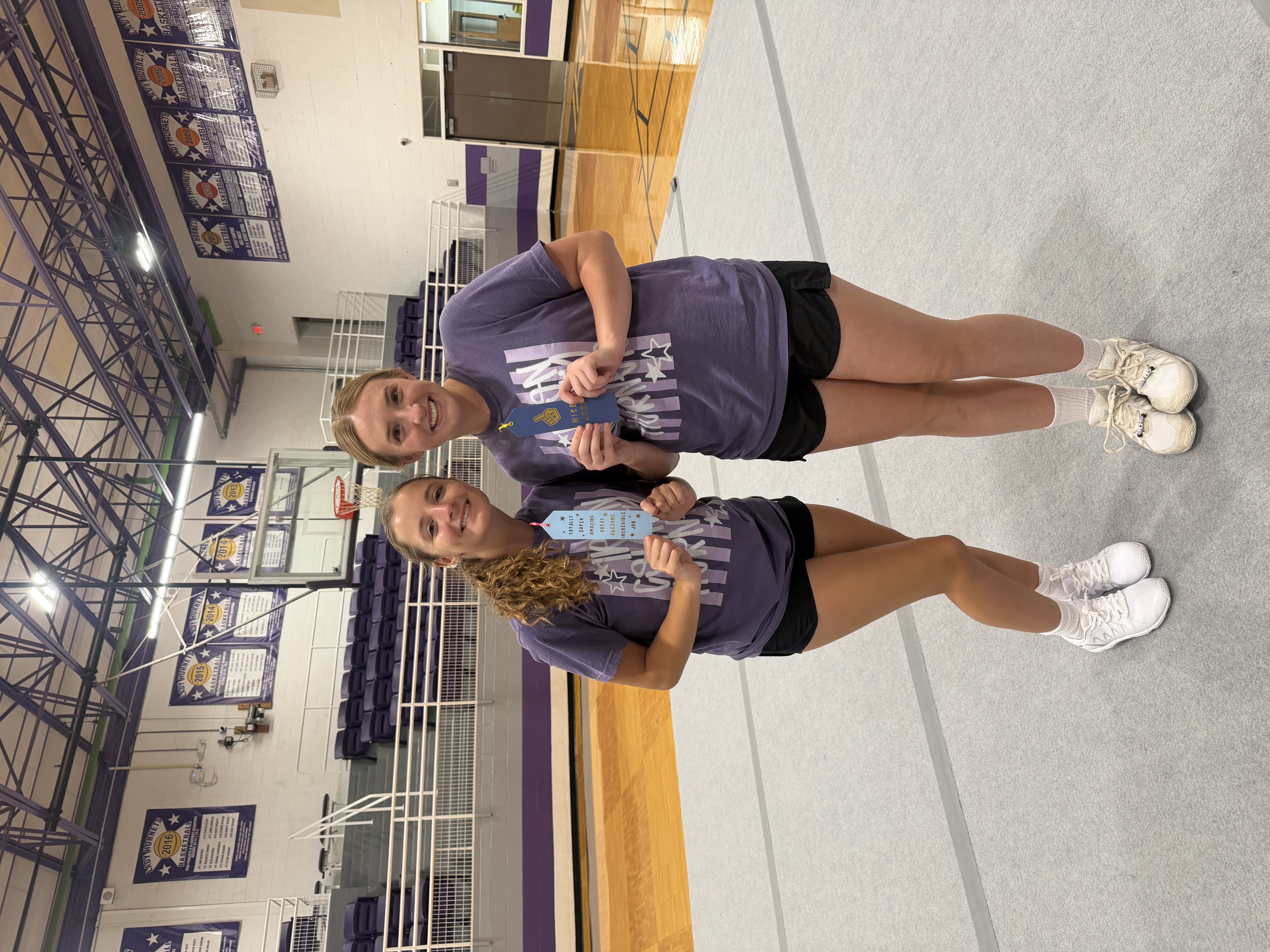 Two smiling young women in purple t-shirts hold blue ribbons in a gymnasium.