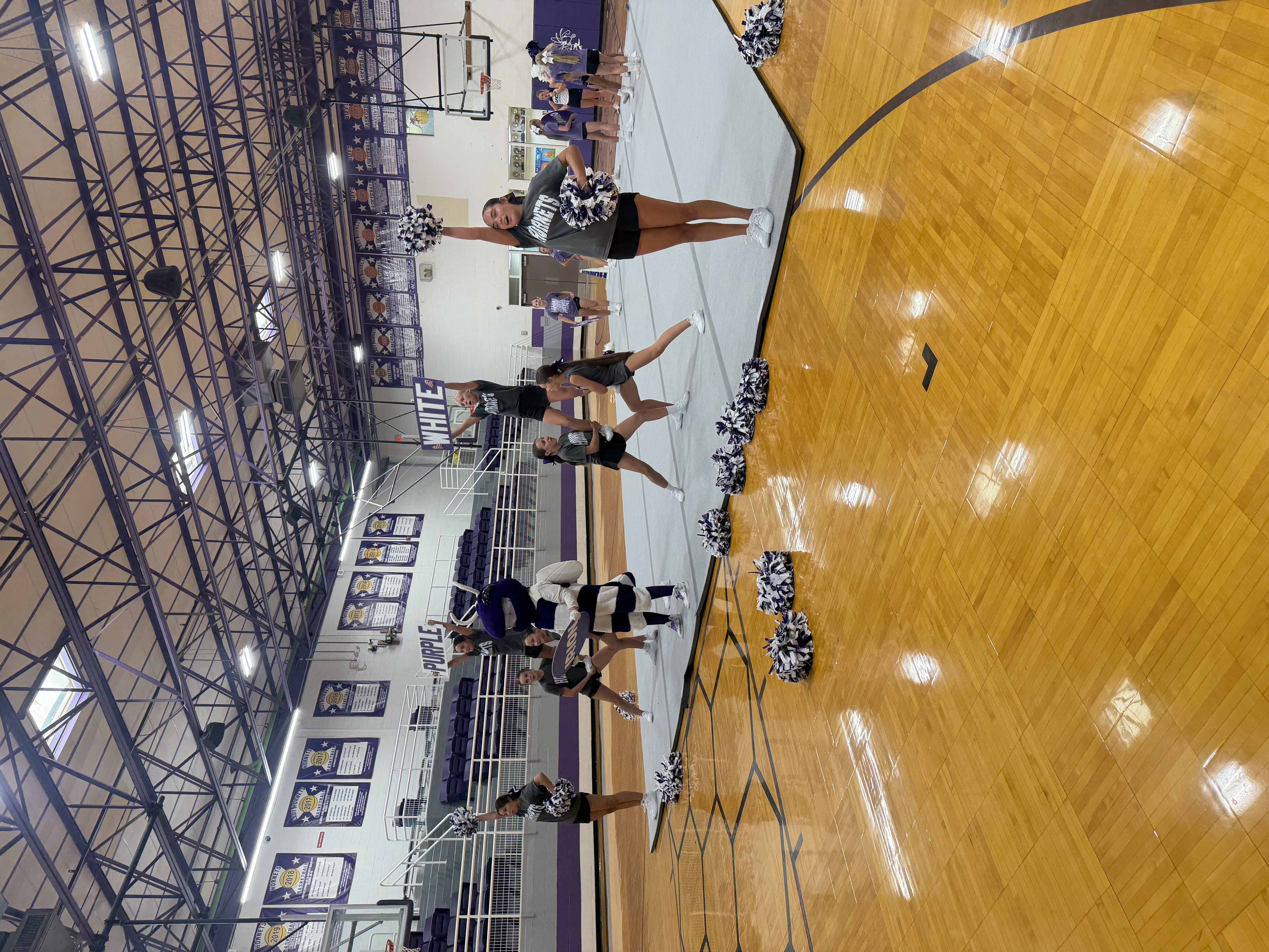 A group of cheerleaders in uniform practice a routine on a basketball court, holding pom-poms.
