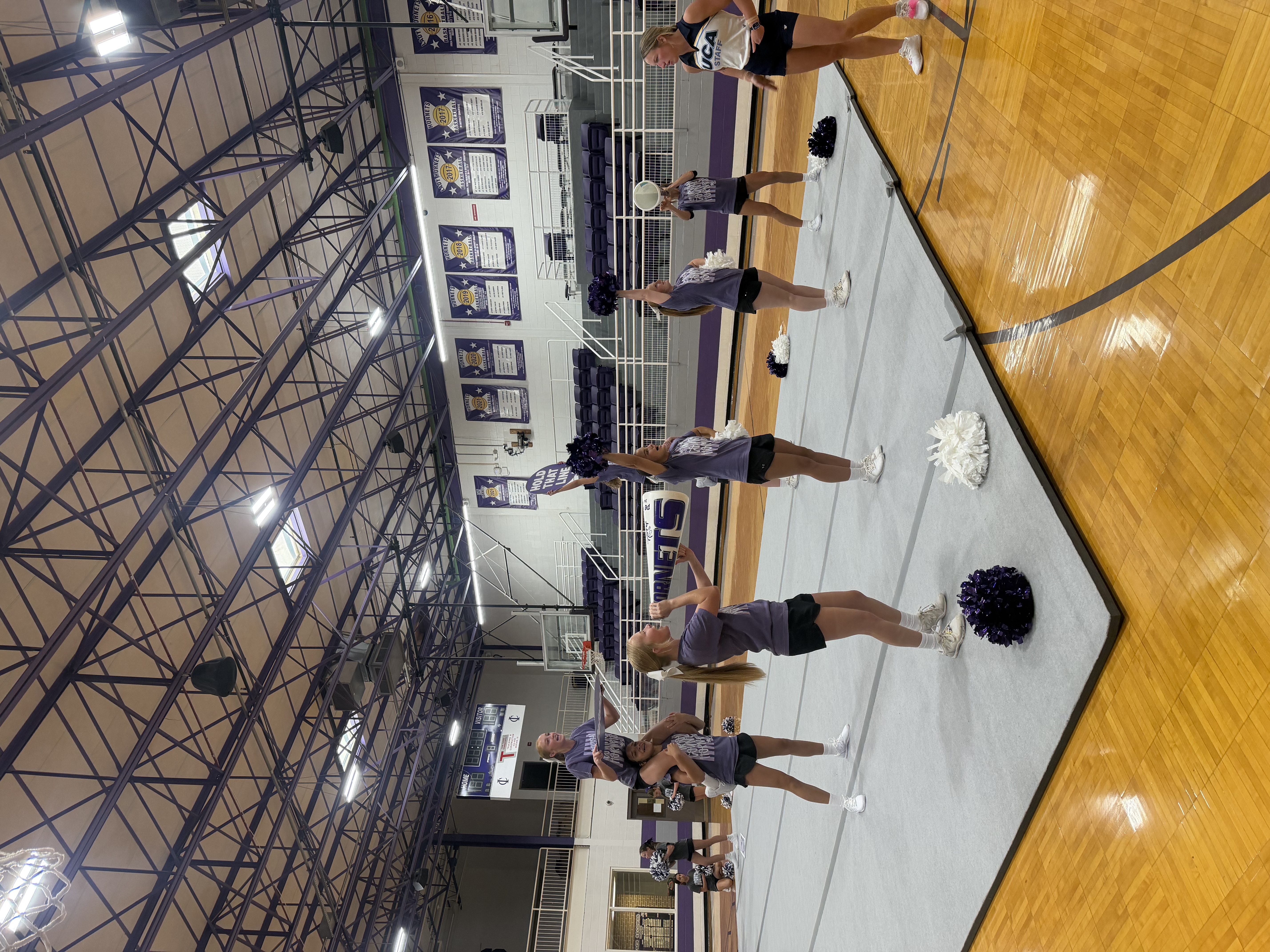 A group of cheerleaders in purple shirts and black shorts practice a routine on a grey mat in a gymnasium.