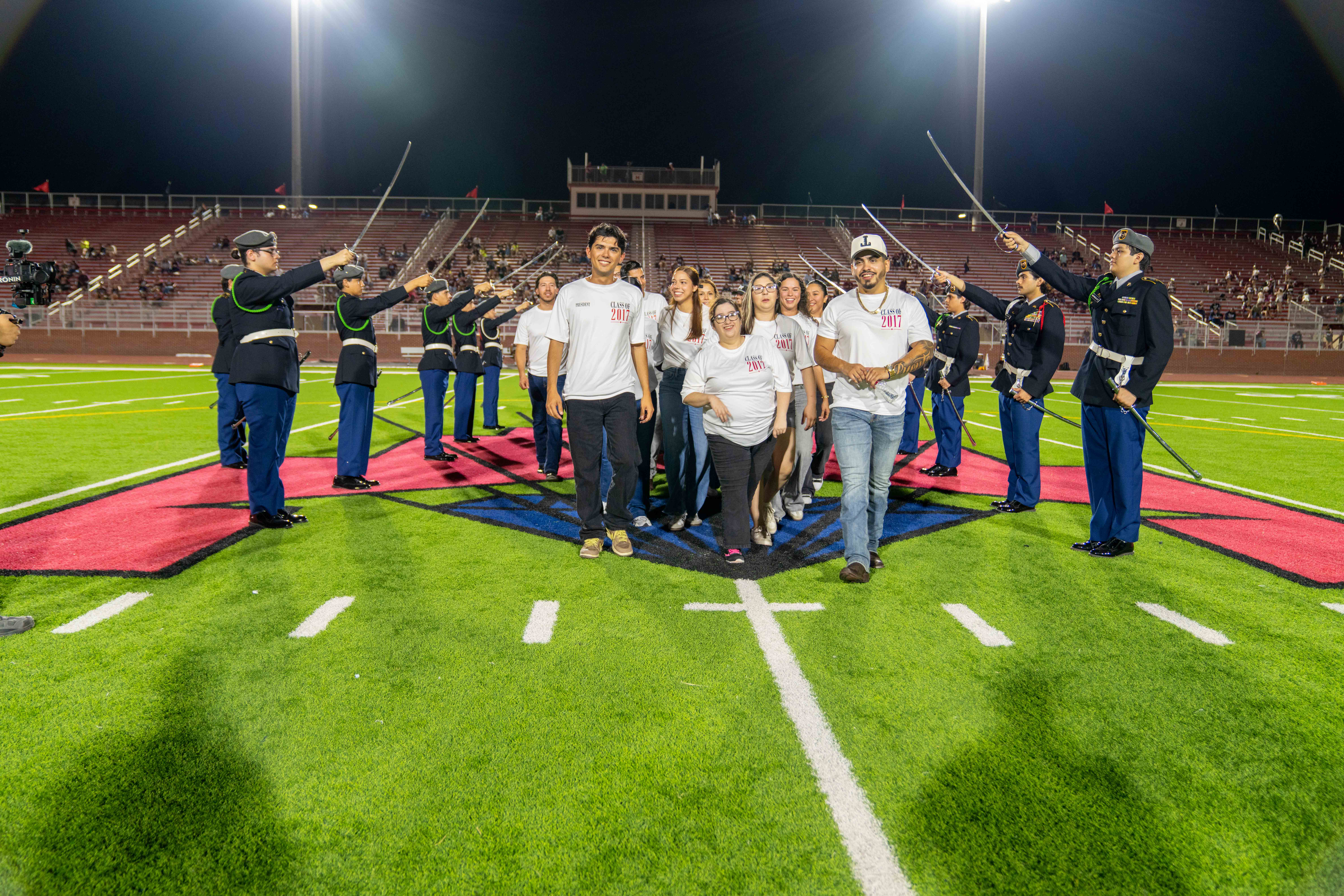 A group of students walking across a lit football field, flanked by a color guard holding raised swords.