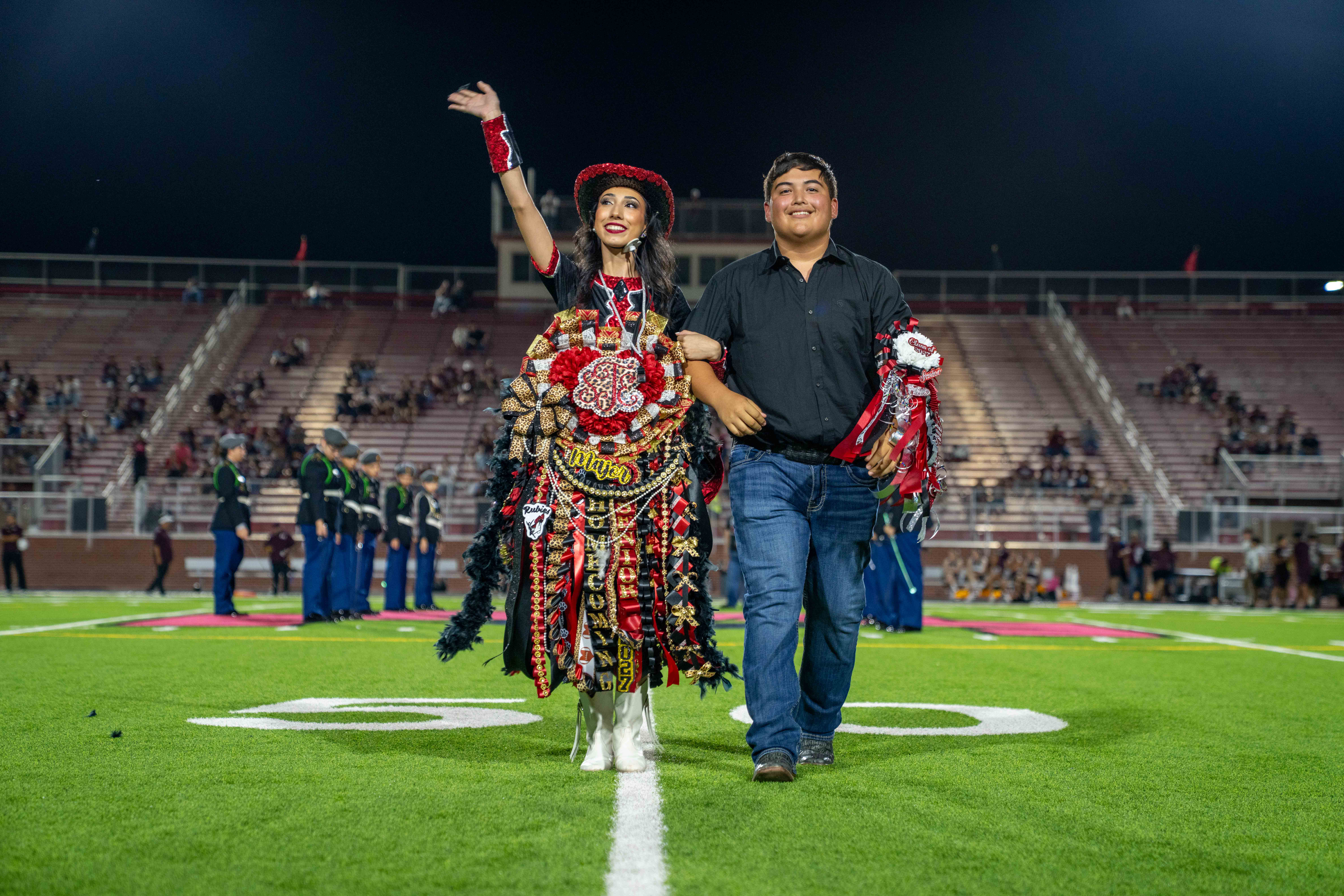 A smiling couple walks arm-in-arm across a glowing football field at night, with the woman waving to the crowd.