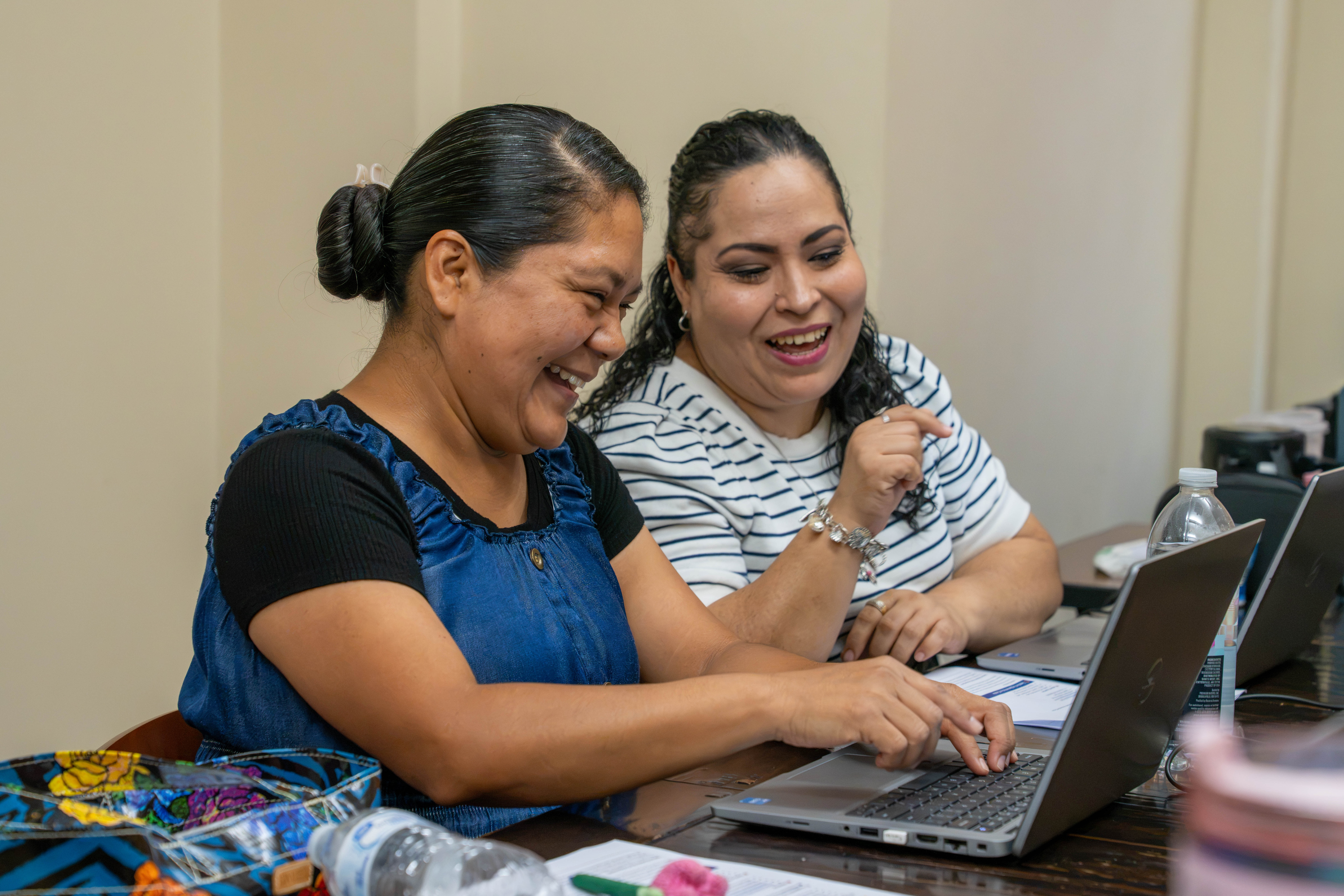 Two women laugh while working together on laptops at a table.