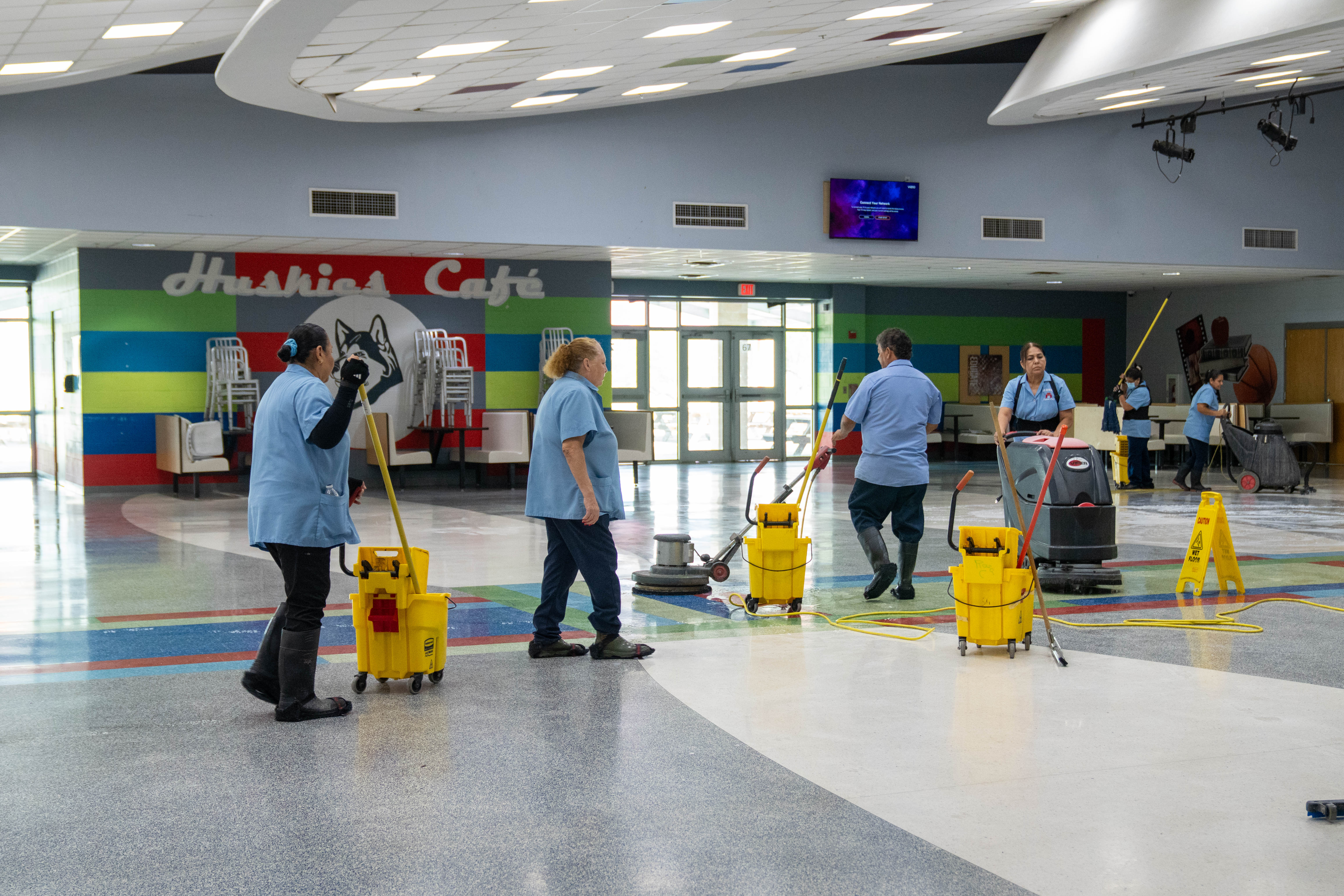 Custodial staff cleans a school hallway with mops and floor scrubbers.