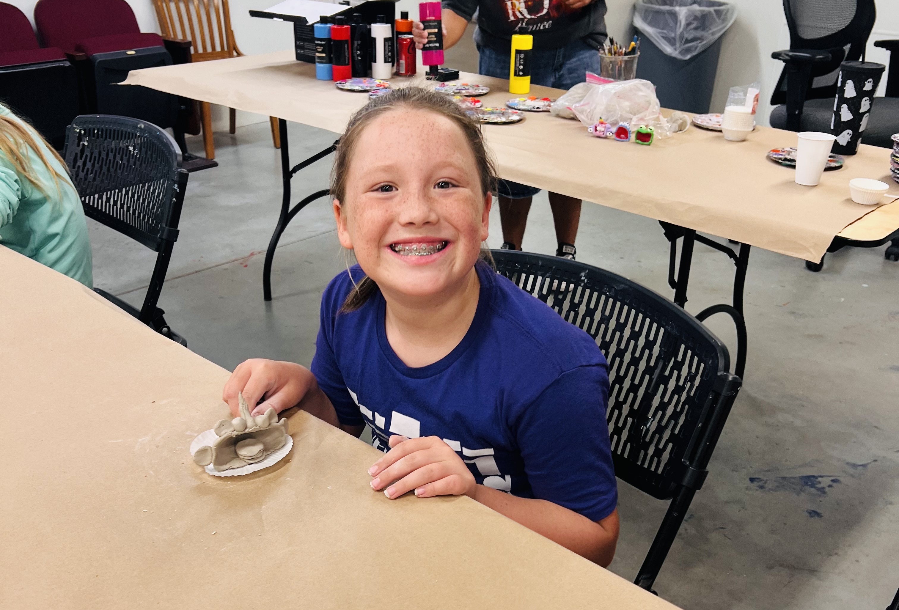 A smiling child with braces sits at a table in a craft room, proudly displaying a small clay figure.