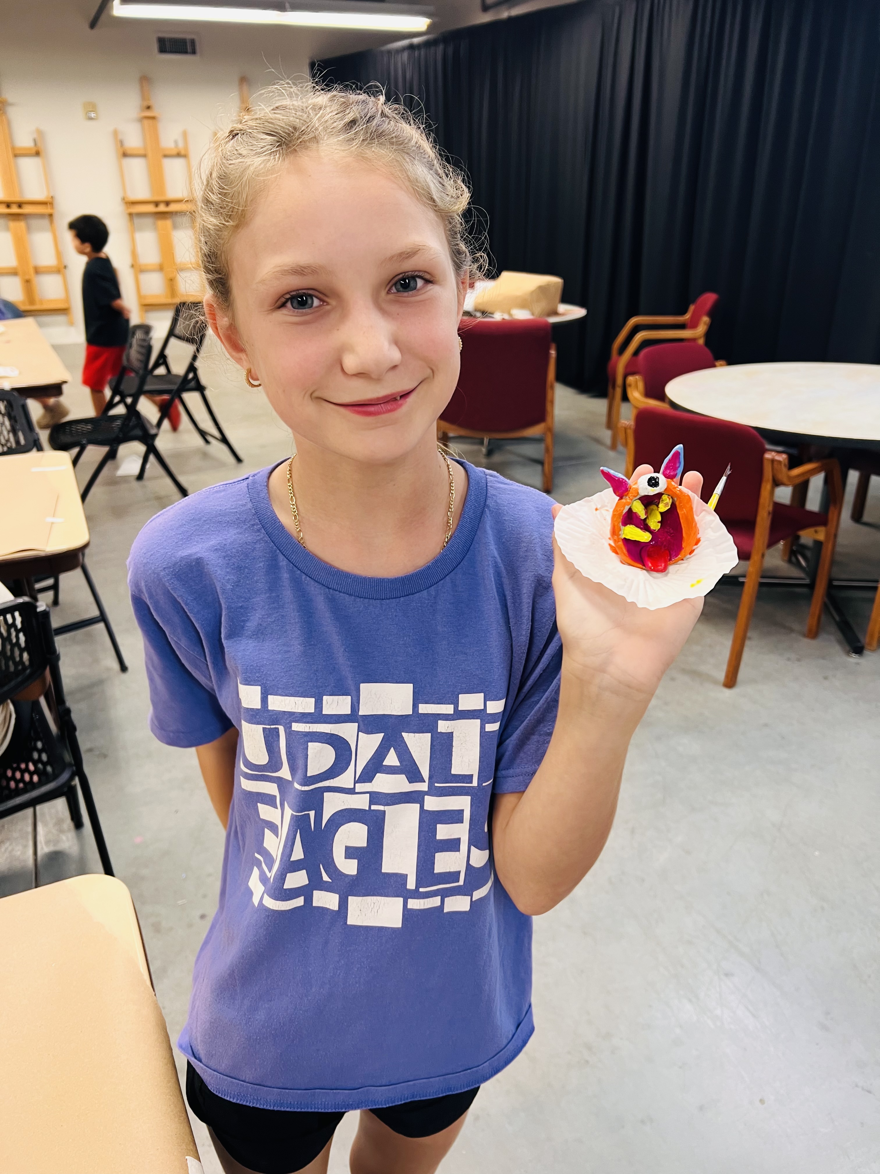 A smiling young girl in an art classroom holds up a small, colorful clay sculpture.