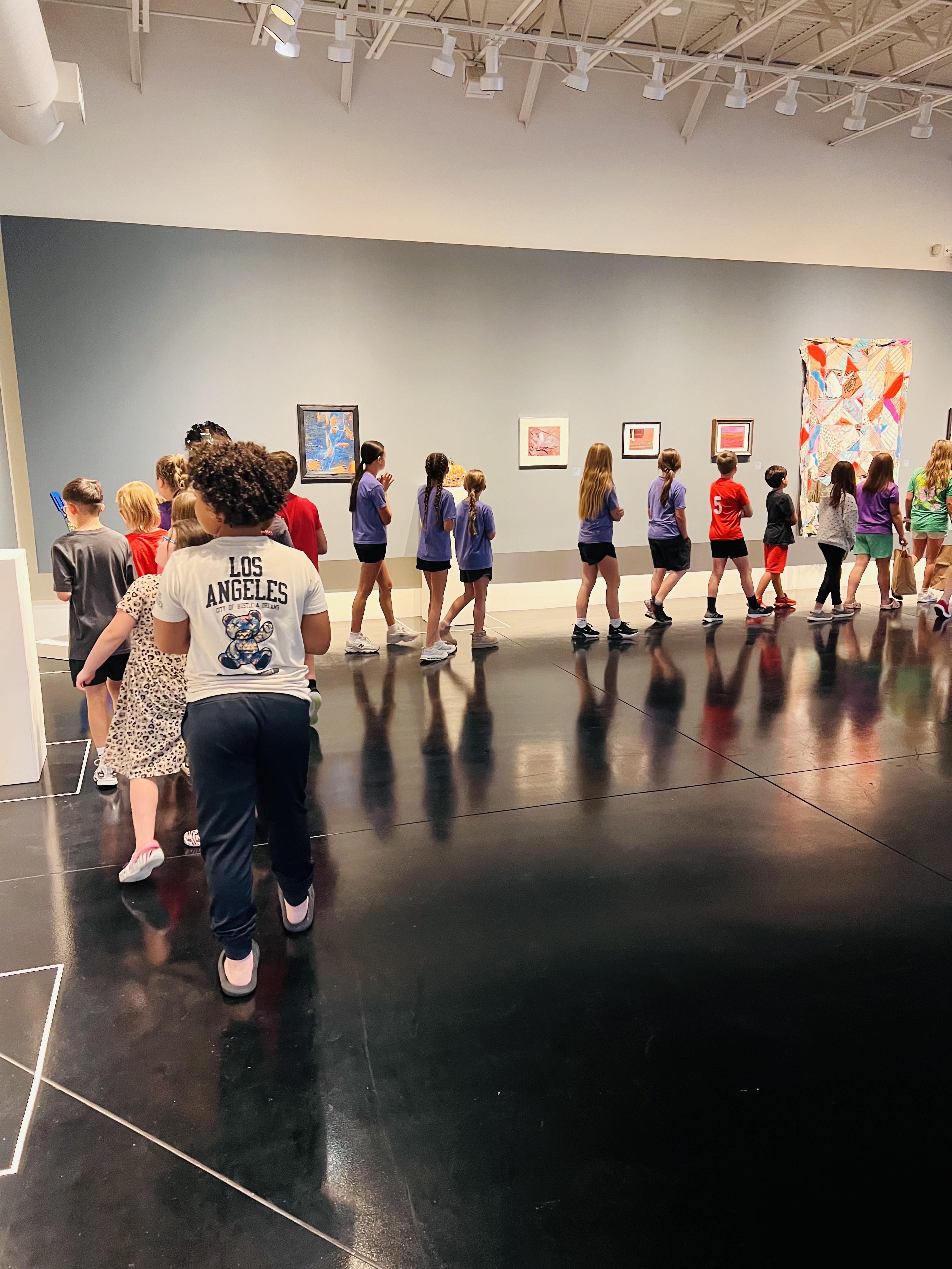 A group of school children walk through an art gallery, viewing paintings and artwork on display along a large gray wall.