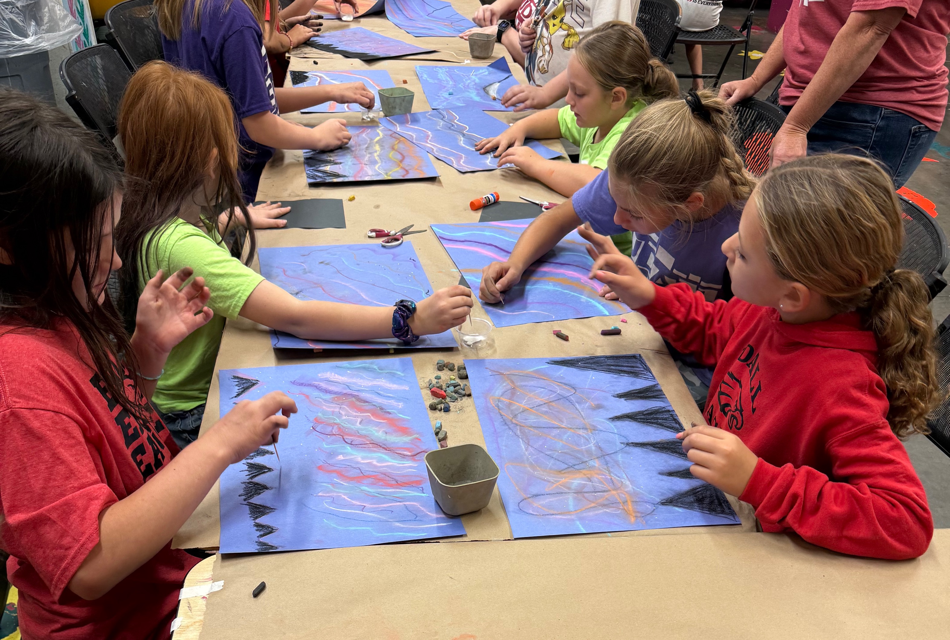 A group of children sits at a long table, working on colorful pastel drawings on blue paper.