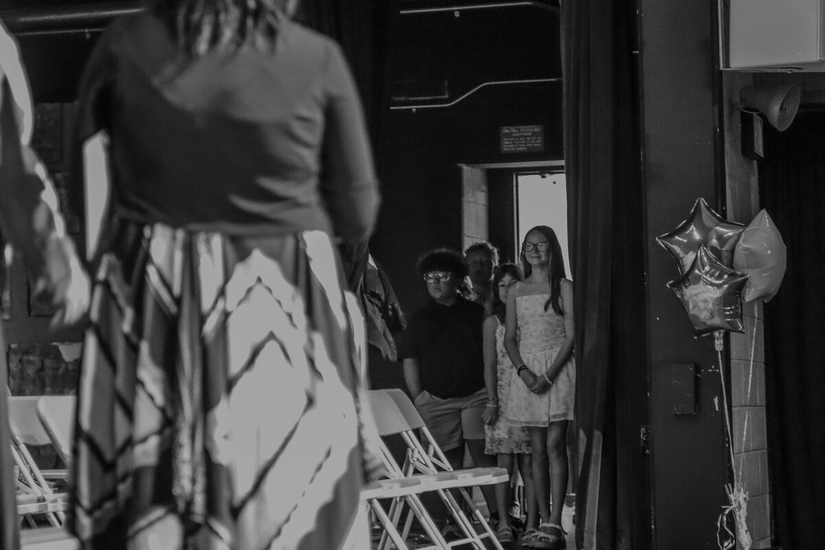 Children stand in a line on a stage, with a woman in the foreground.