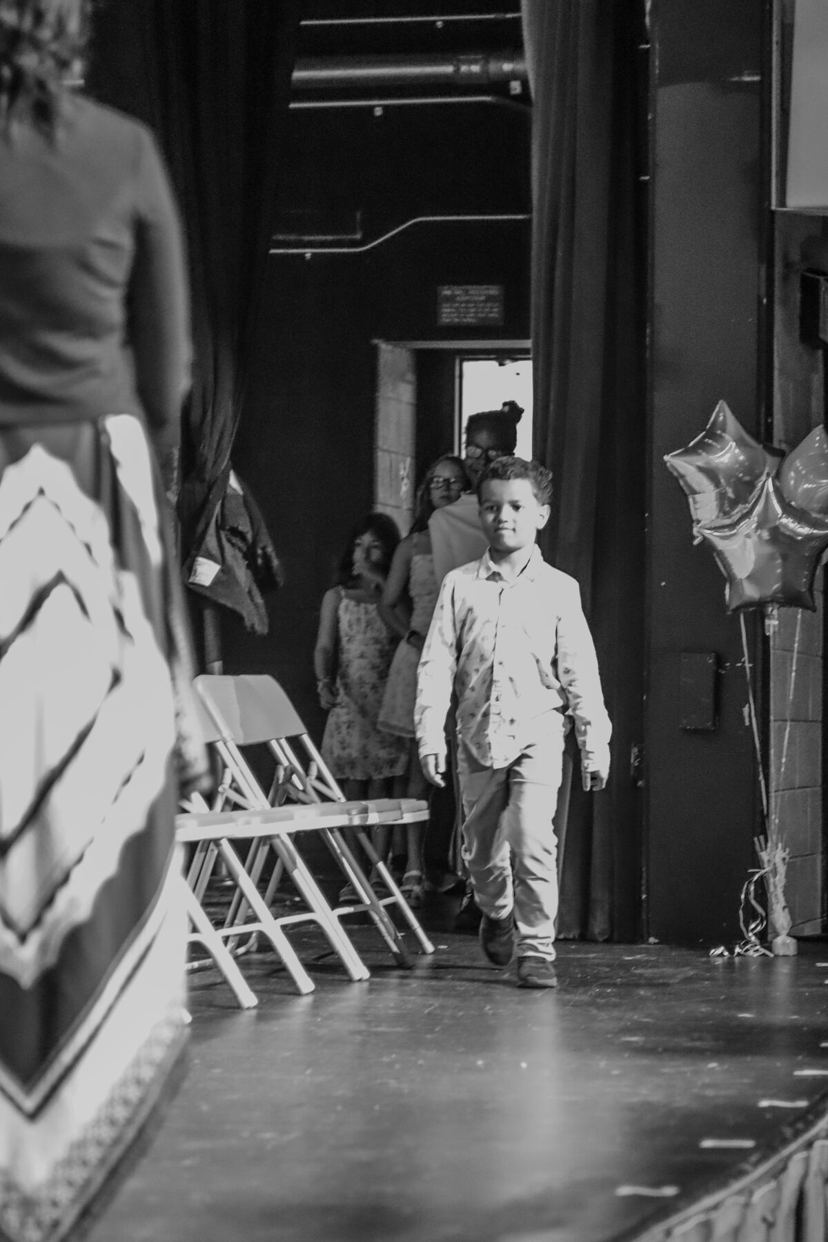 A young boy in a patterned shirt walks confidently across a stage, with other children and chairs in the background.