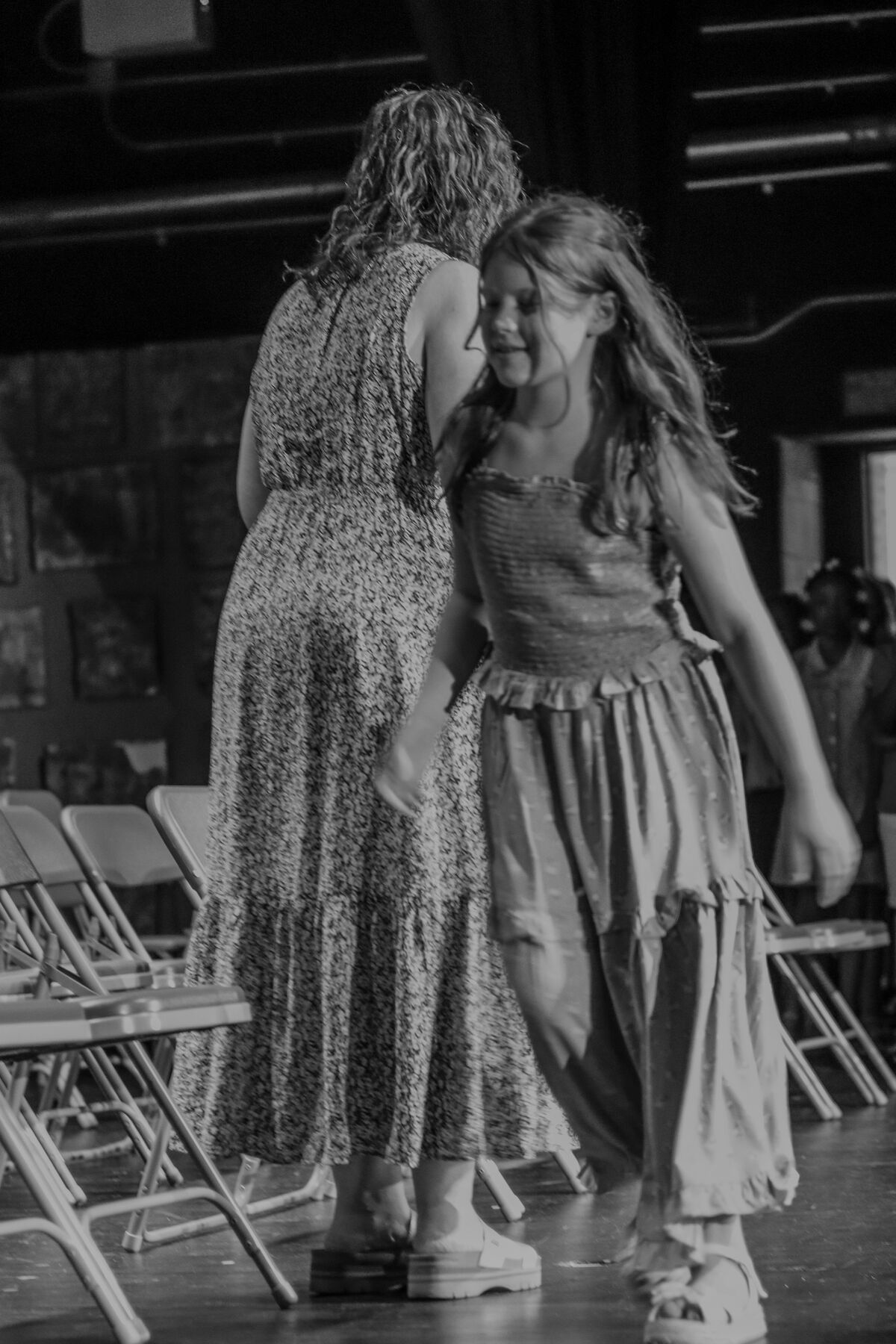 A young girl in a ruffled dress smiles as she walks past a woman in a patterned dress.
