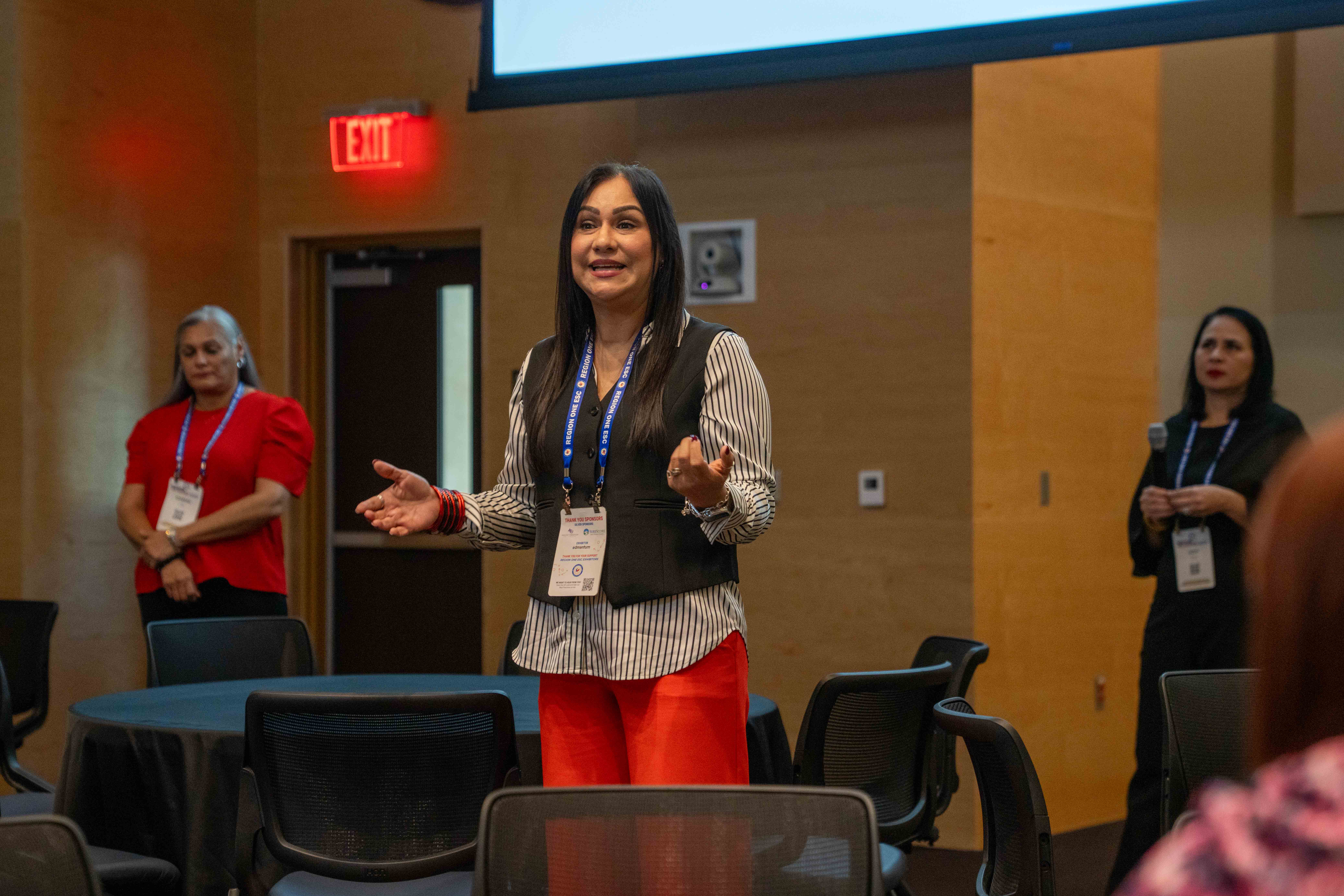 A speaker with long dark hair gestures while presenting at an indoor event, with attendees visible in the background.