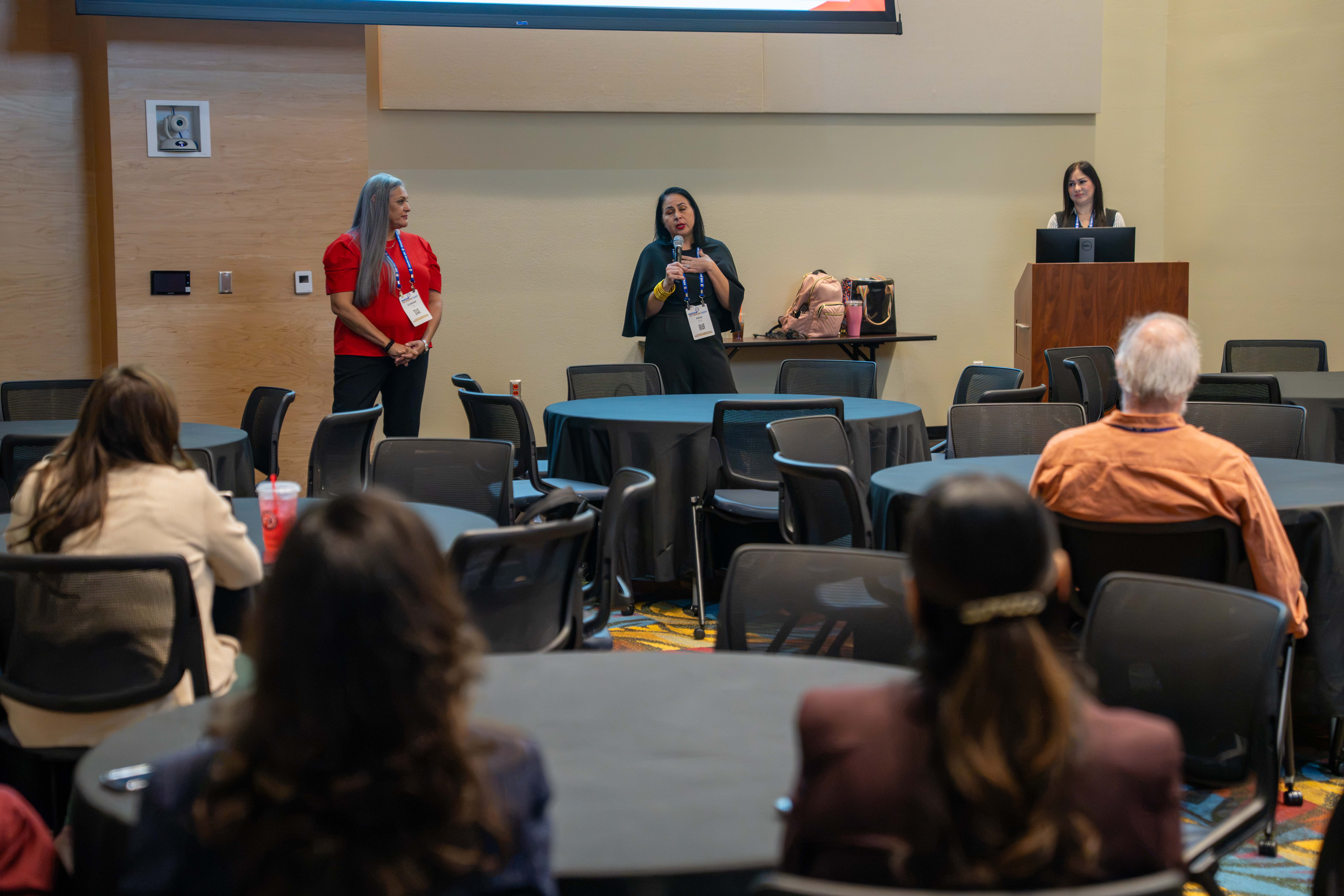 A woman speaks into a microphone at the front of a conference room while another woman stands beside her, facing an audience seated at round tables.