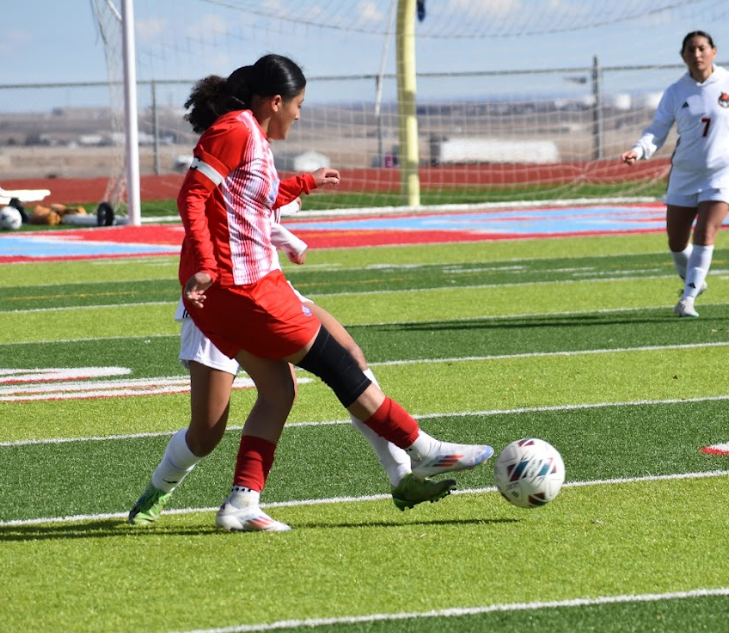 A soccer player in a red and white striped jersey kicks a soccer ball on a green turf field.