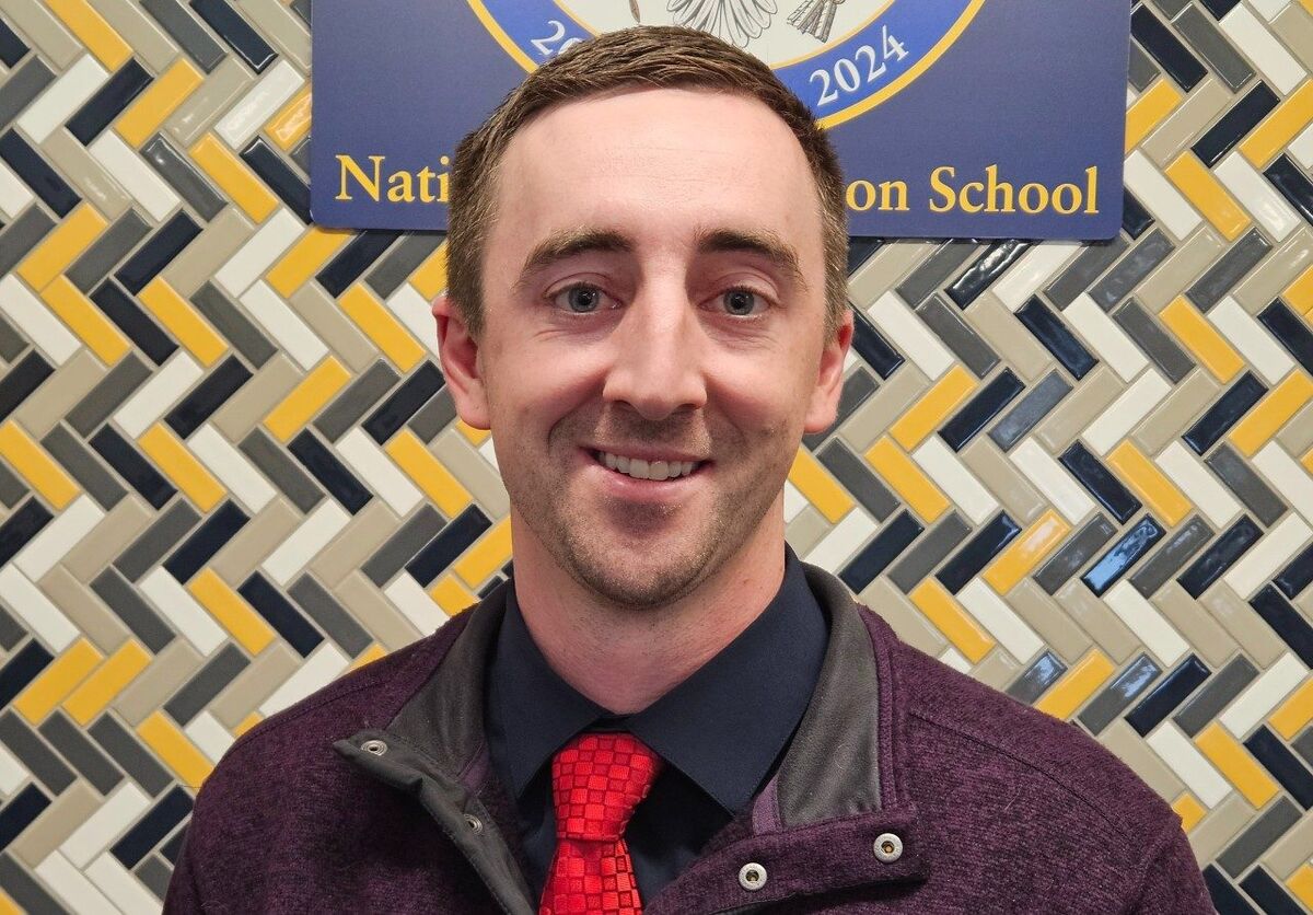 A man in a dark shirt and red tie smiles in front of a colorful tiled wall.