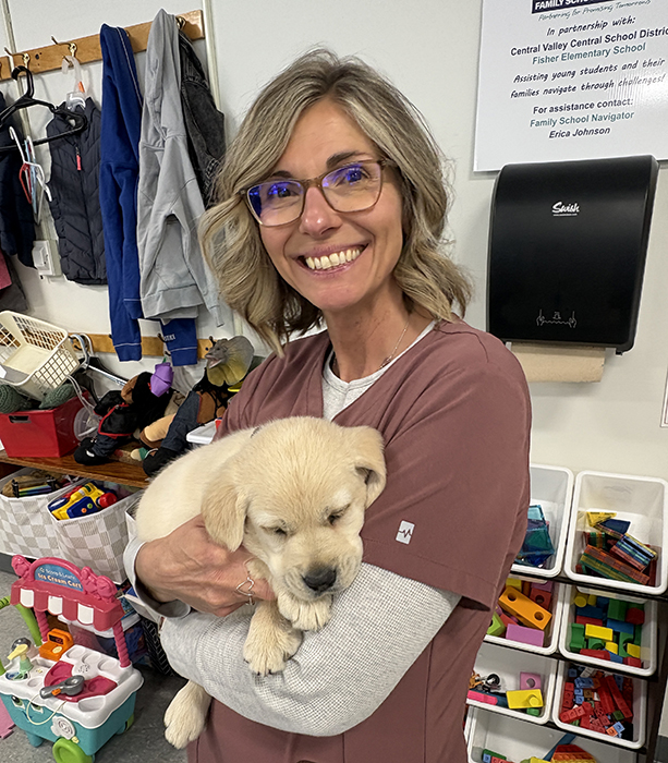 A smiling woman in glasses holds a sleeping yellow Labrador puppy in a room with children's toys and coats.