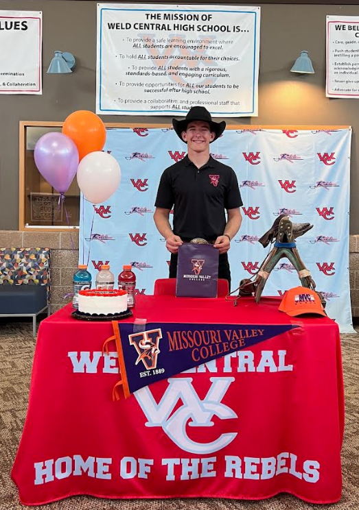 A young man in a cowboy hat smiles while holding a bag from Missouri Valley College, standing at a table with a cake and balloons.