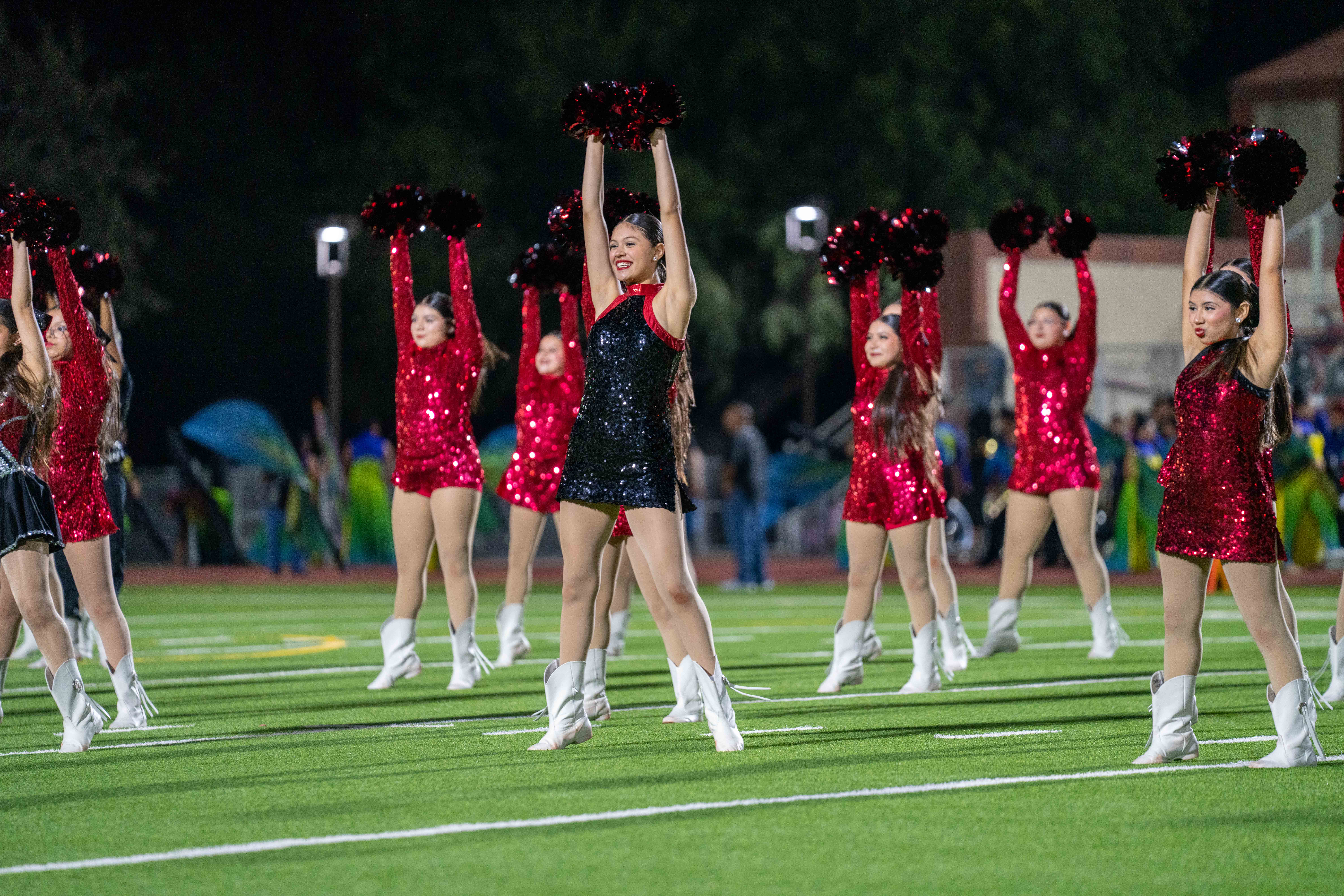 A line of drill team members in red and black sequined uniforms perform on a football field at night.