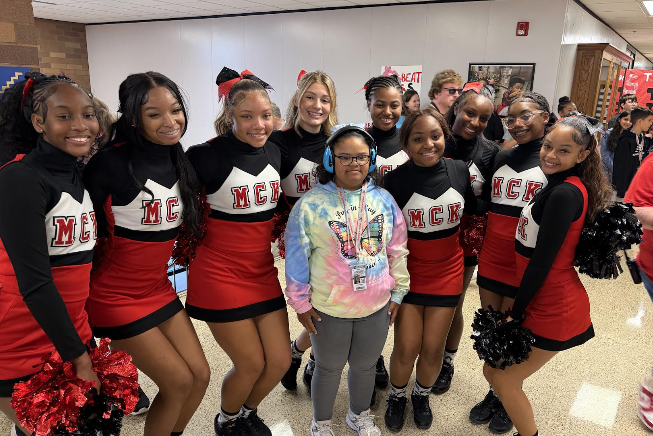 A group of cheerleaders in red, black, and white uniforms pose together in a school hallway.