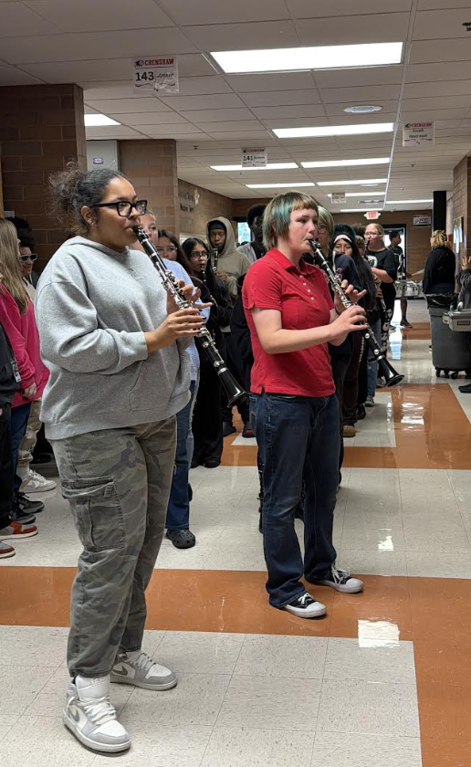 Two students practice playing clarinets in a school hallway, with other students and band members standing in line behind them.
