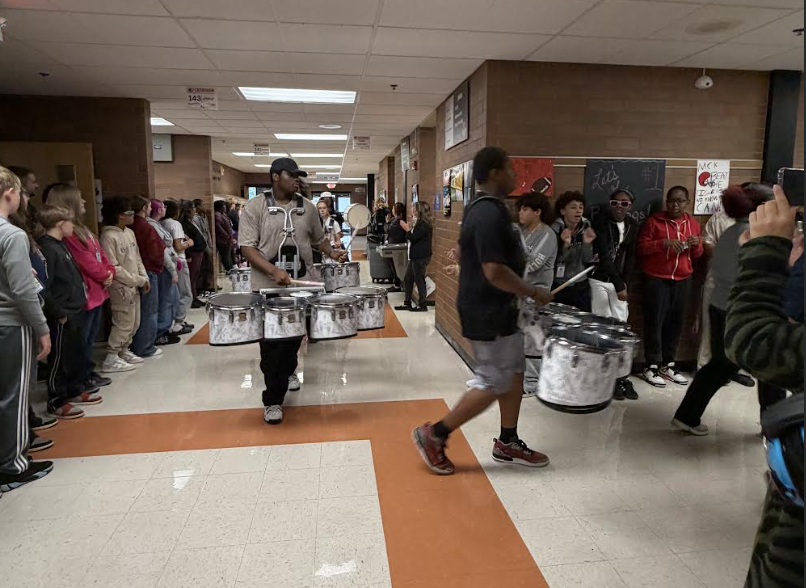 Two student drummers march down a school hallway, carrying quad drums while other students watch.