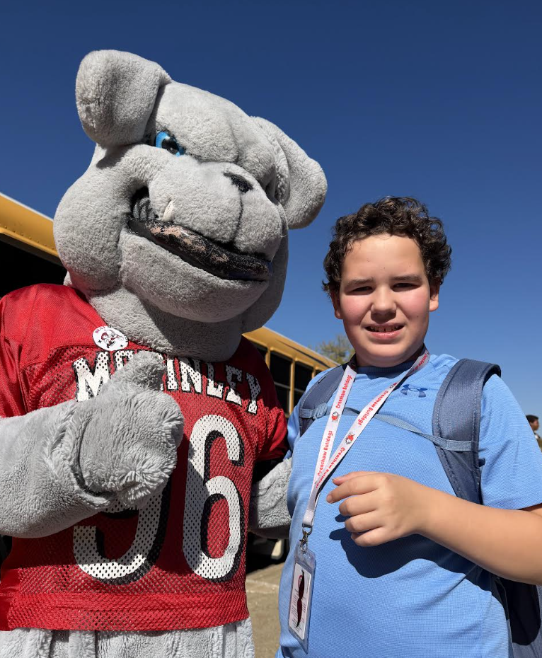 A smiling boy poses outdoors next to a large grey bulldog mascot wearing a red jersey.