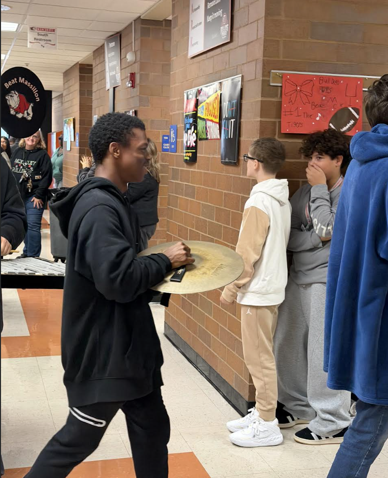 A student in a black hoodie carries a brass cymbal down a school hallway while three other students watch.