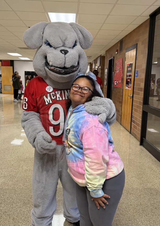A smiling student hugs a grey bulldog mascot in a school hallway.