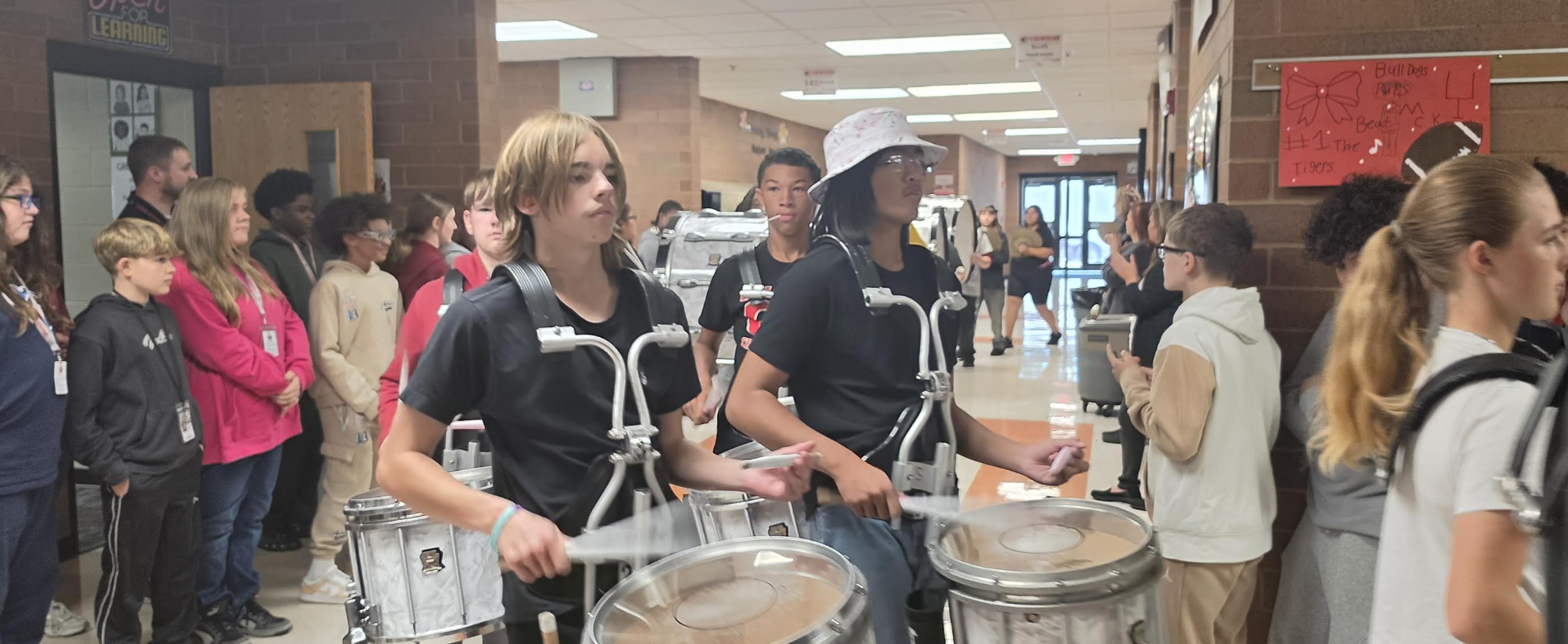 A marching band drumline walks through a brightly lit school hallway carrying drums.
